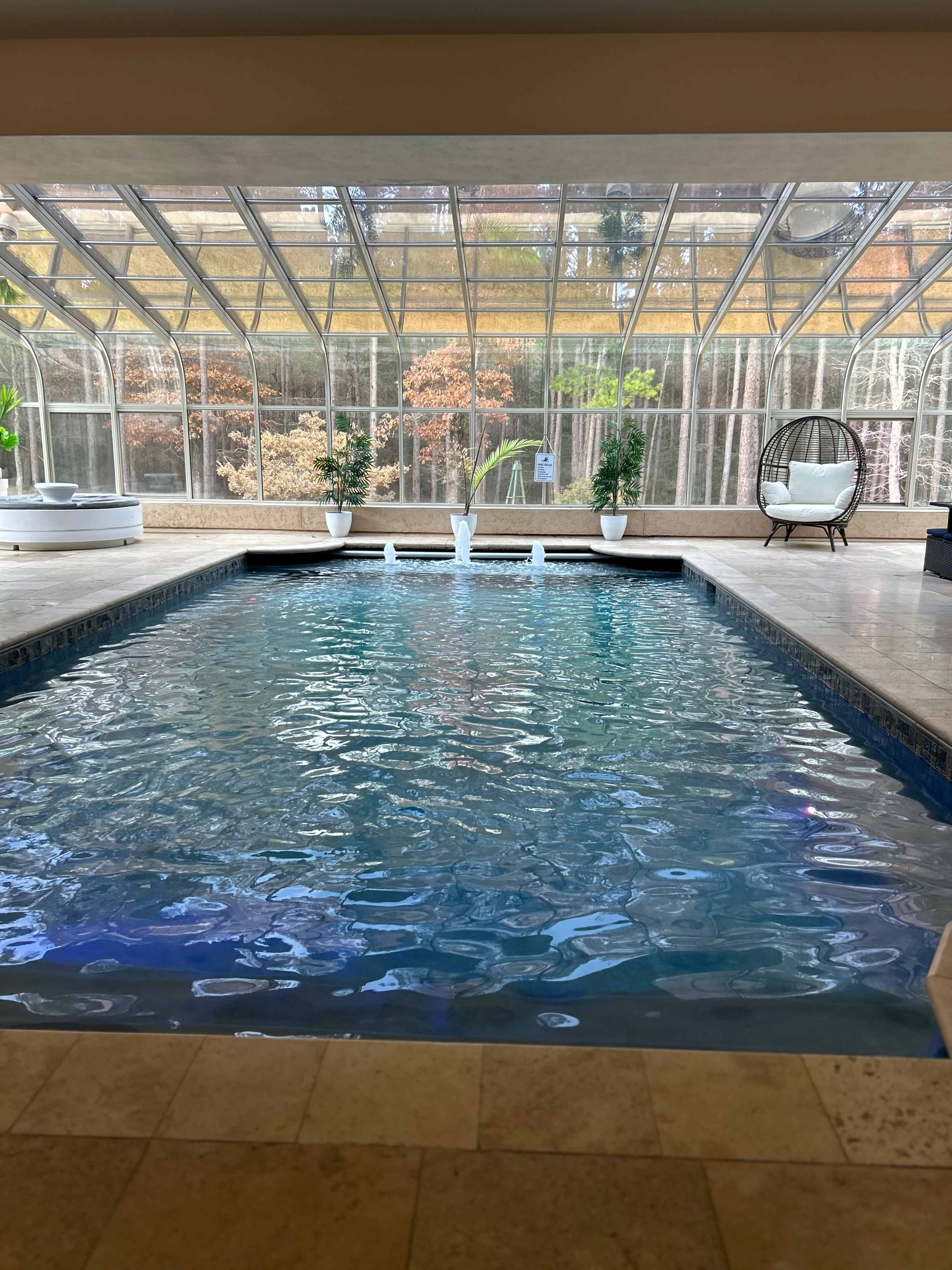 An indoor swimming pool enclosed by a glass structure, surrounded by potted plants and a lounge chair.
