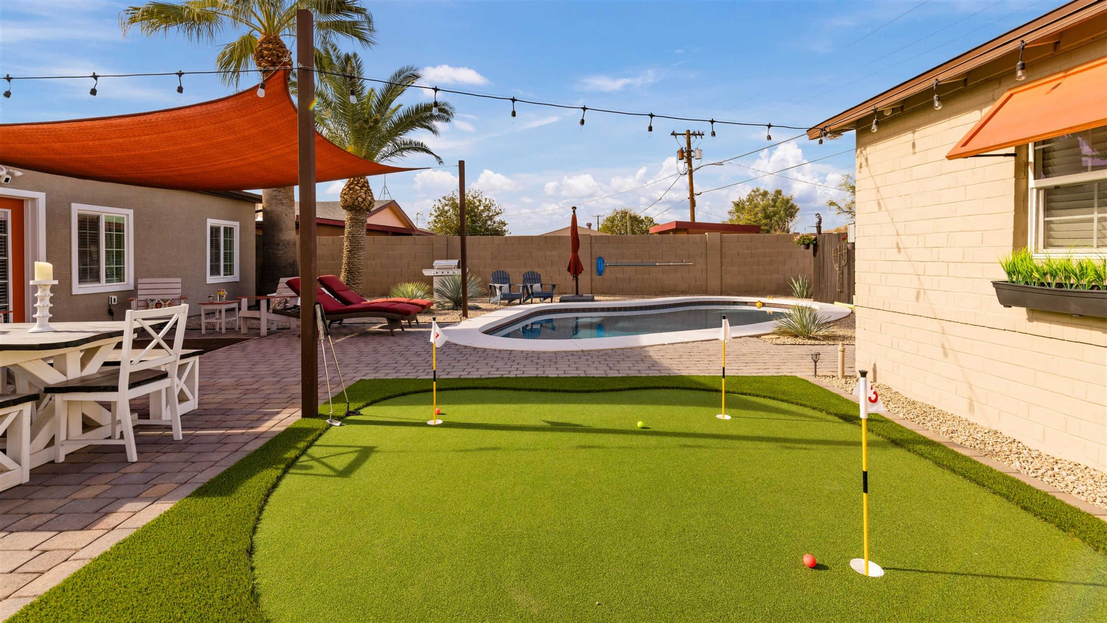 A backyard featuring a putting green, a swimming pool, lounge chairs, and a patio area with a dining table.