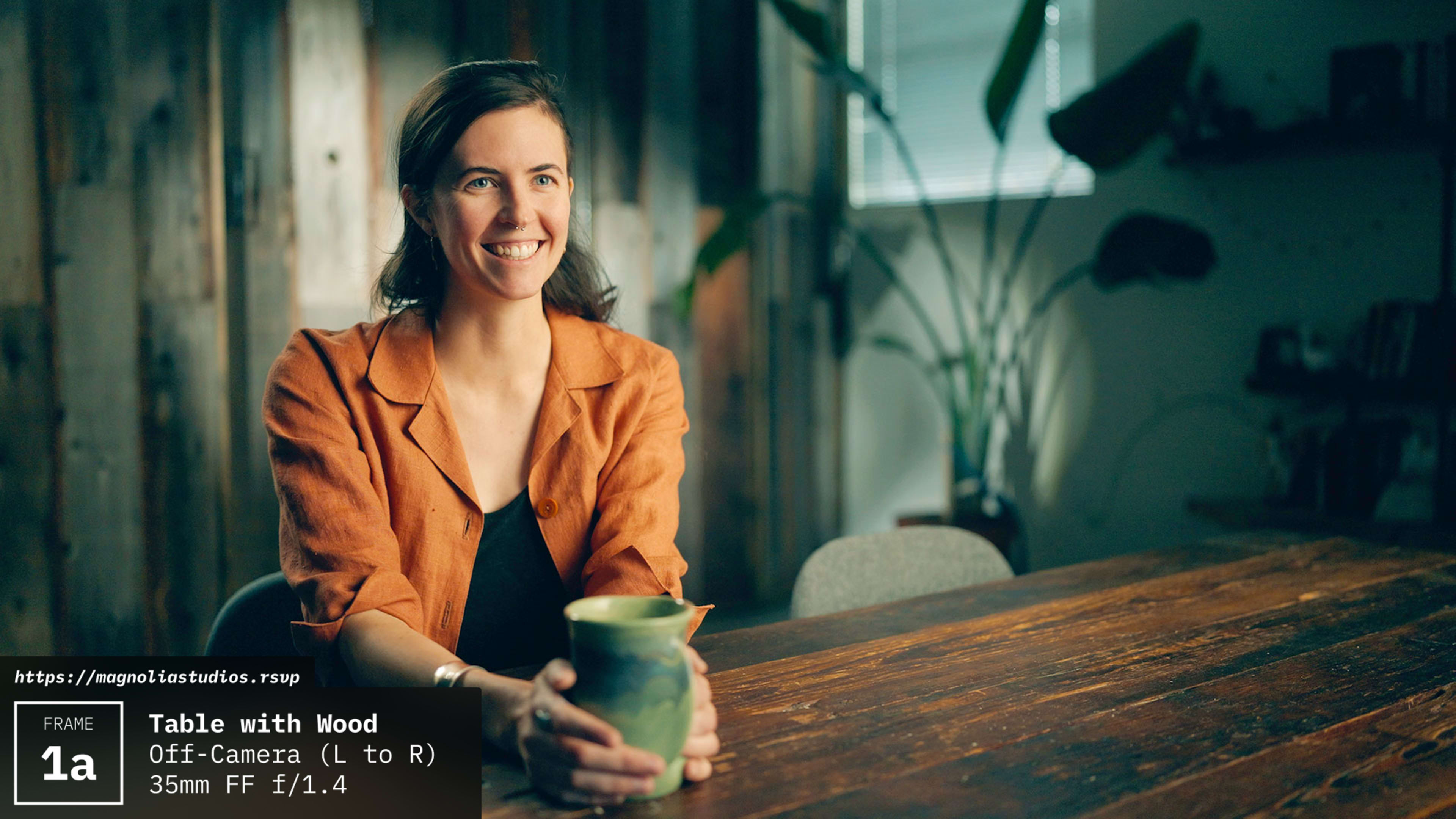 A woman with long hair sits at a table made of wood, holding a ceramic mug, with plants and wooden panels in the background.
