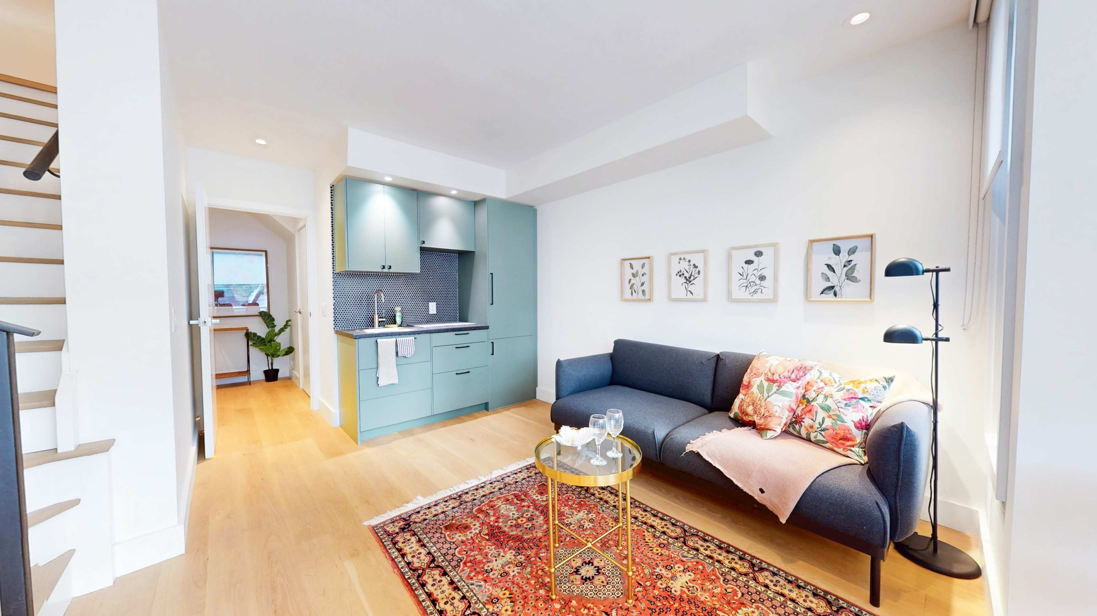 The image shows a modern living space featuring a blue sofa, a small round table with two glasses, a kitchenette with teal cabinets, and decorative art on the walls.