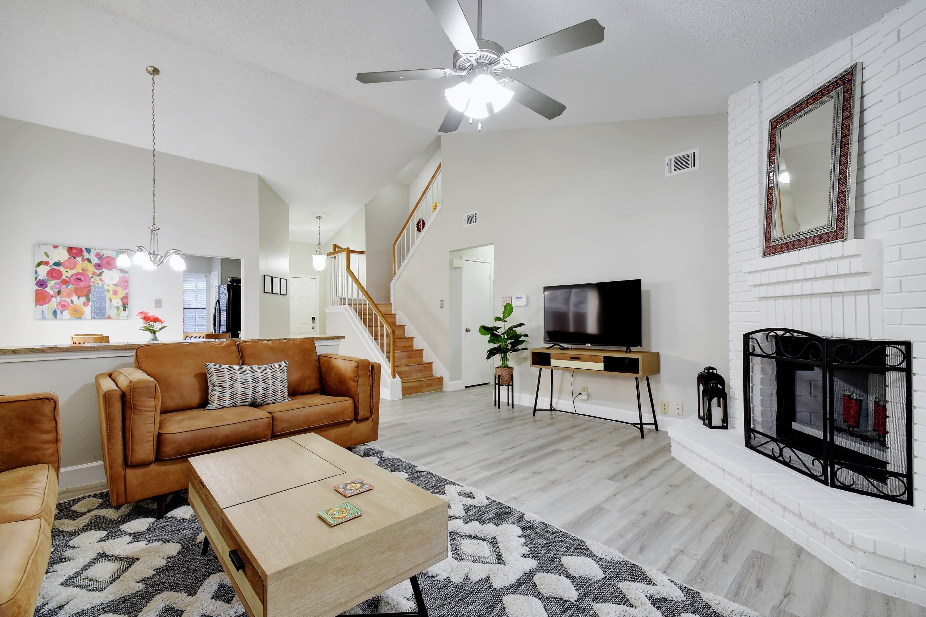 The image shows a spacious living room featuring a brown leather sofa, a coffee table, a television on a stand, and a fireplace, with stairs visible in the background.