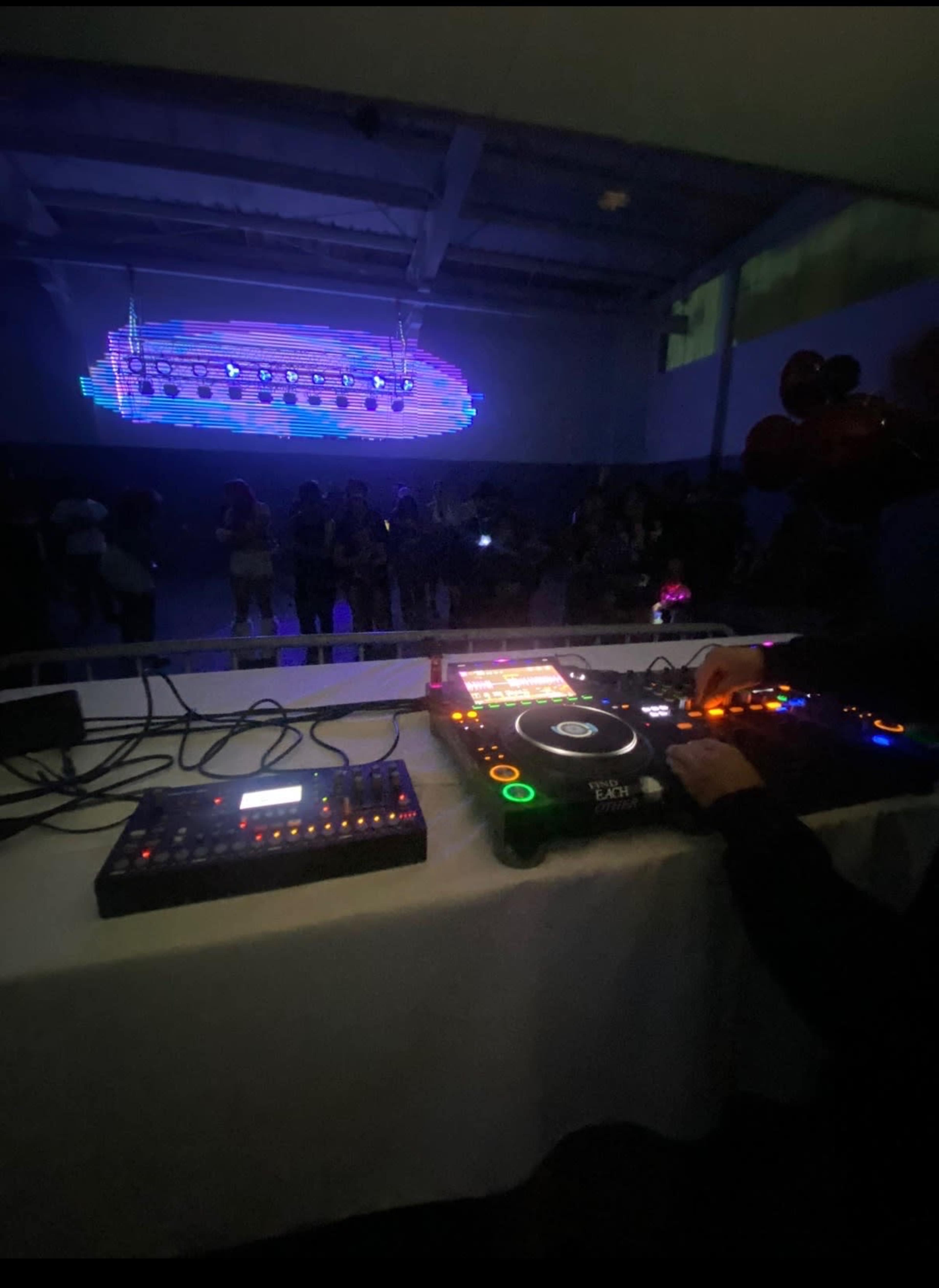 A DJ operates turntables and equipment in front of a crowd illuminated by colorful lights in a dimly lit venue.