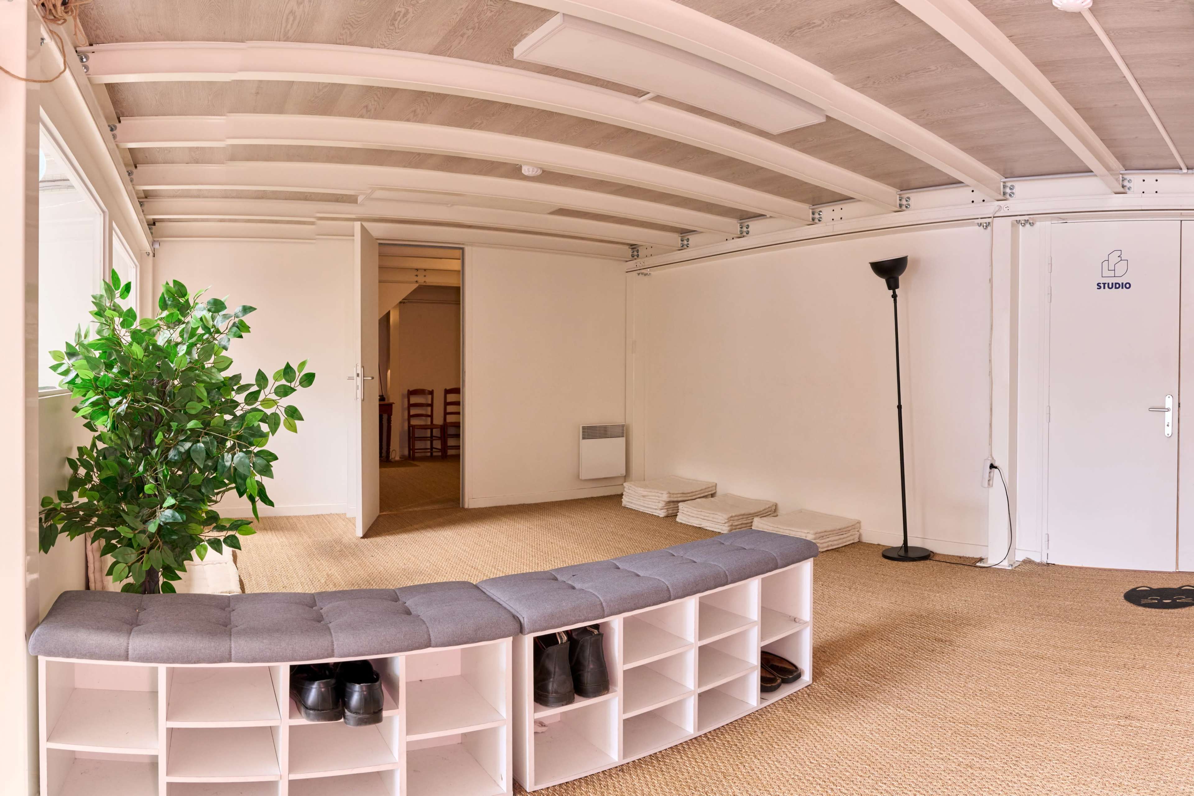 The image shows a minimalist interior space featuring a bench with shoe storage, a potted plant, a standing lamp, and a door labeled "STUDIO."