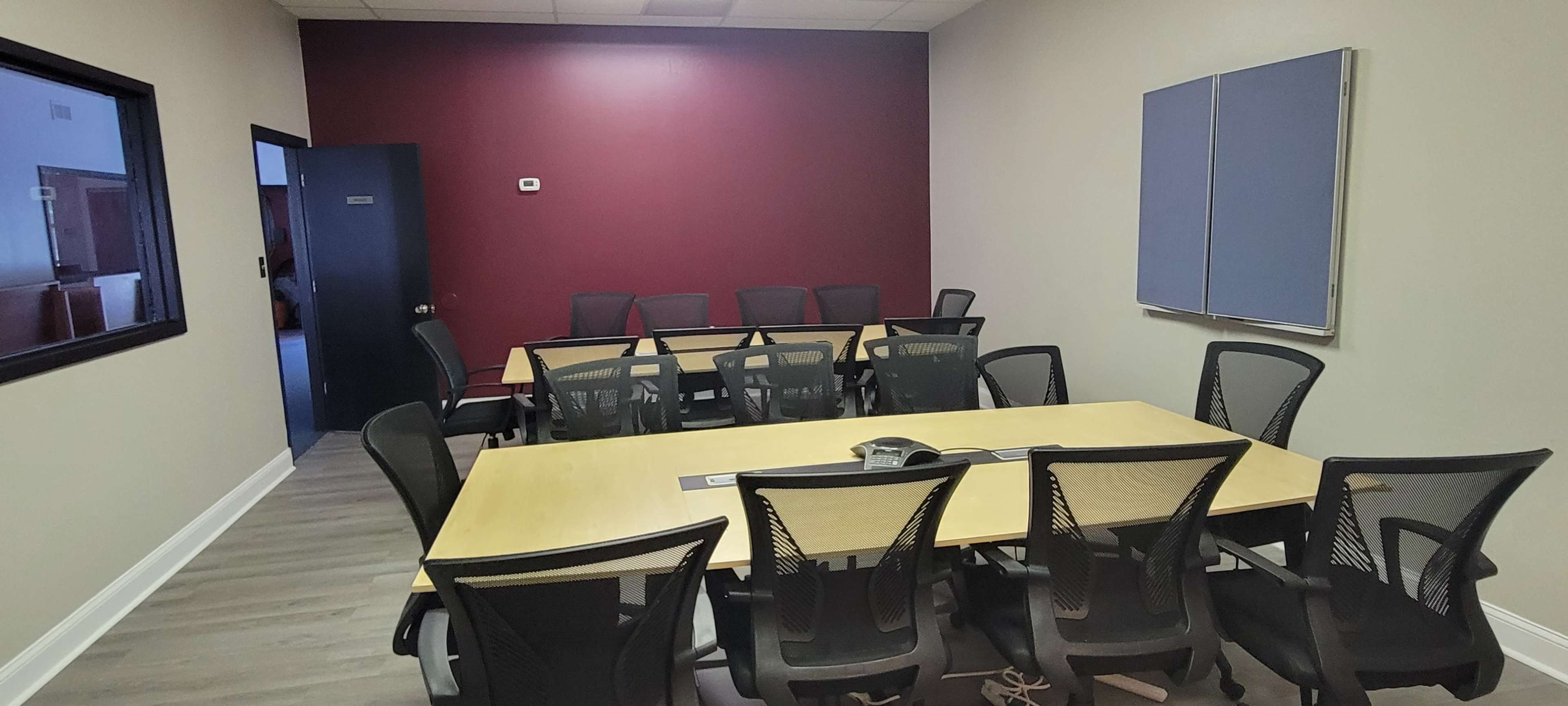 A conference room with a long wooden table surrounded by black mesh chairs, a purple accent wall, and a blue presentation board.