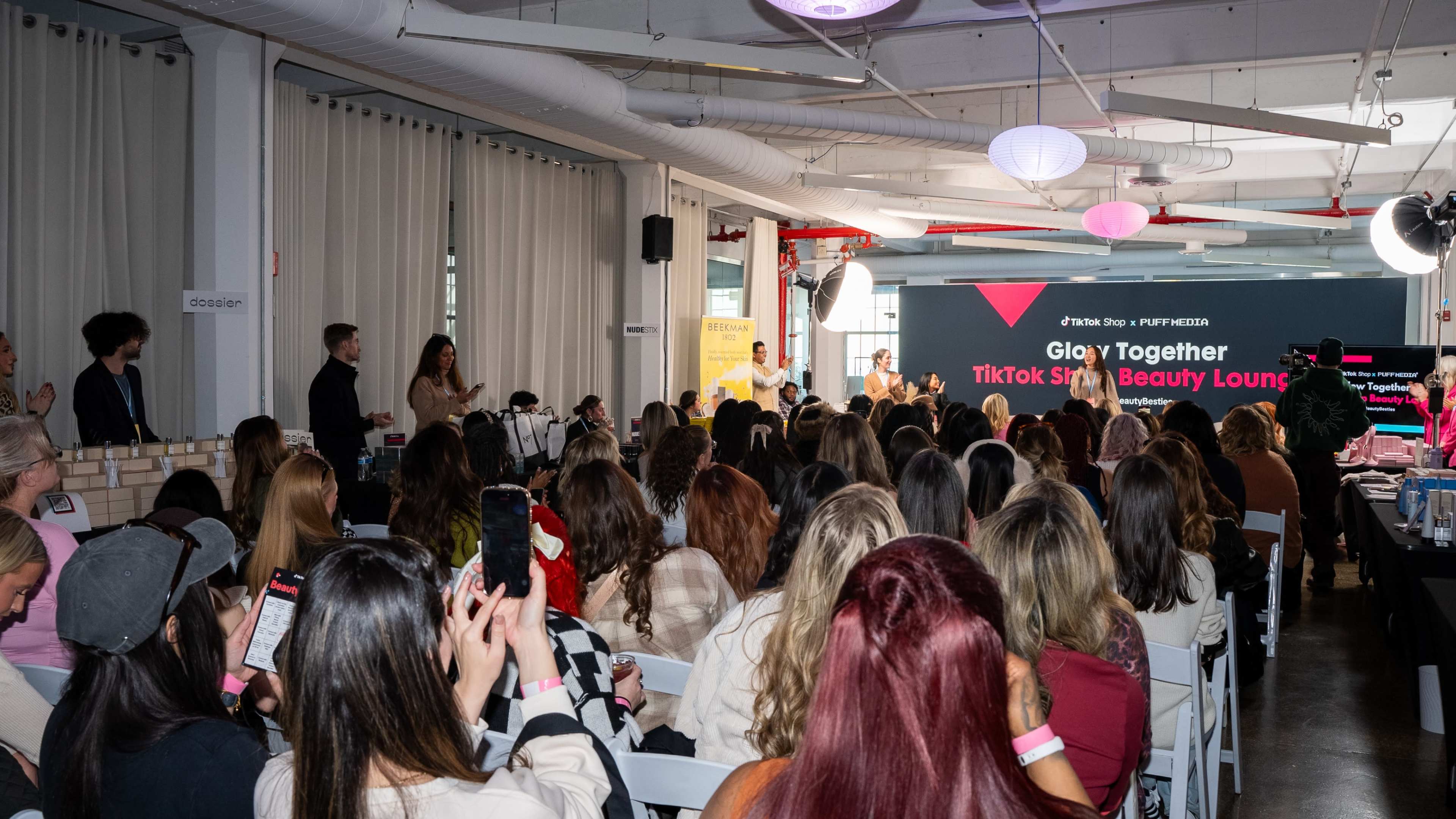 A large audience is seated in a well-lit venue, watching a presentation or event titled "Glow Together TikTok Style Beauty Lounge."