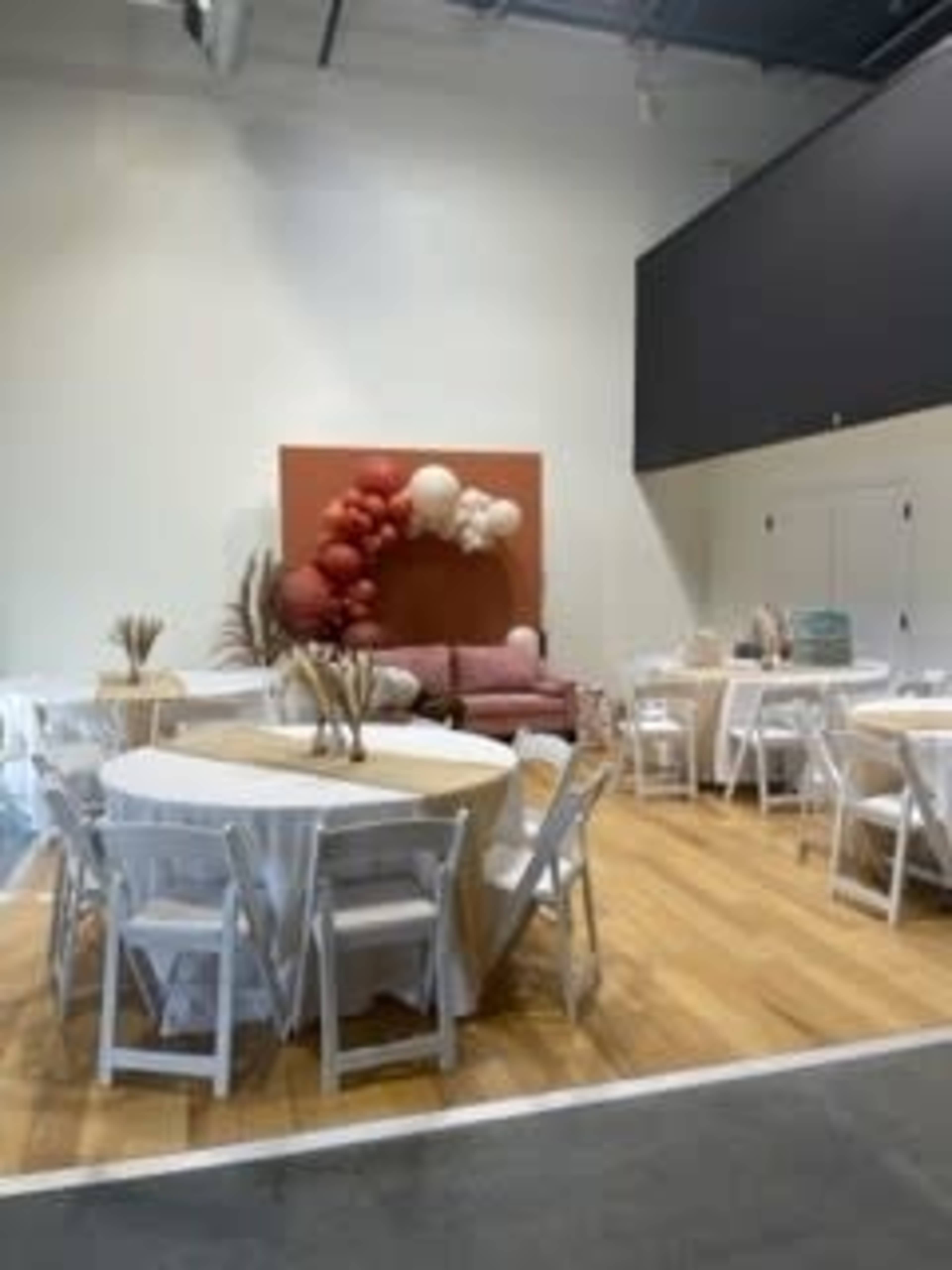 A spacious event venue with several round tables set up, white chairs, and a decorative wall featuring balloons and a couch in the background.
