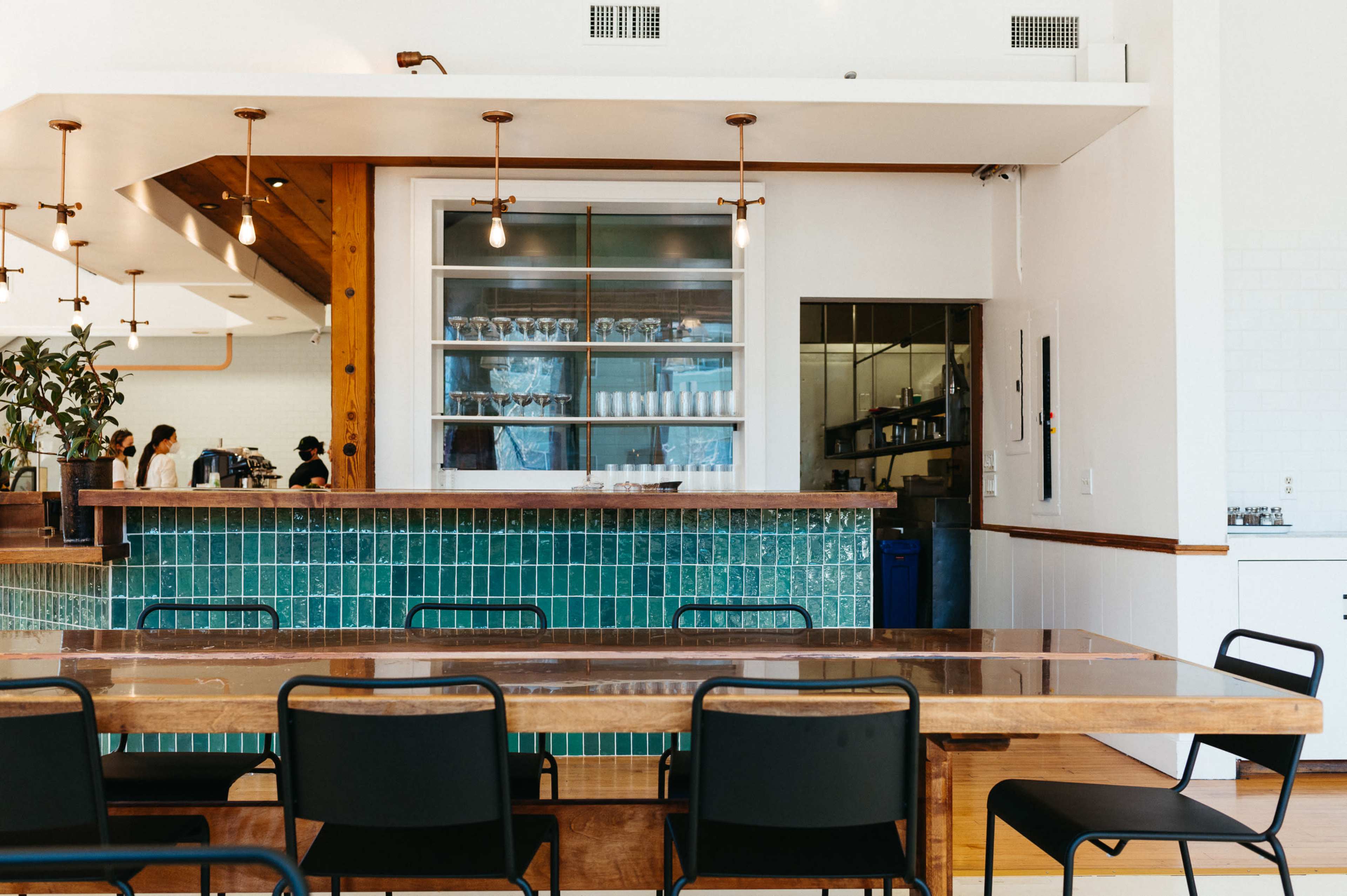 A modern café interior features a wooden counter with teal tiles, a large window showcasing glassware, and simple black chairs around a sizeable wooden table.