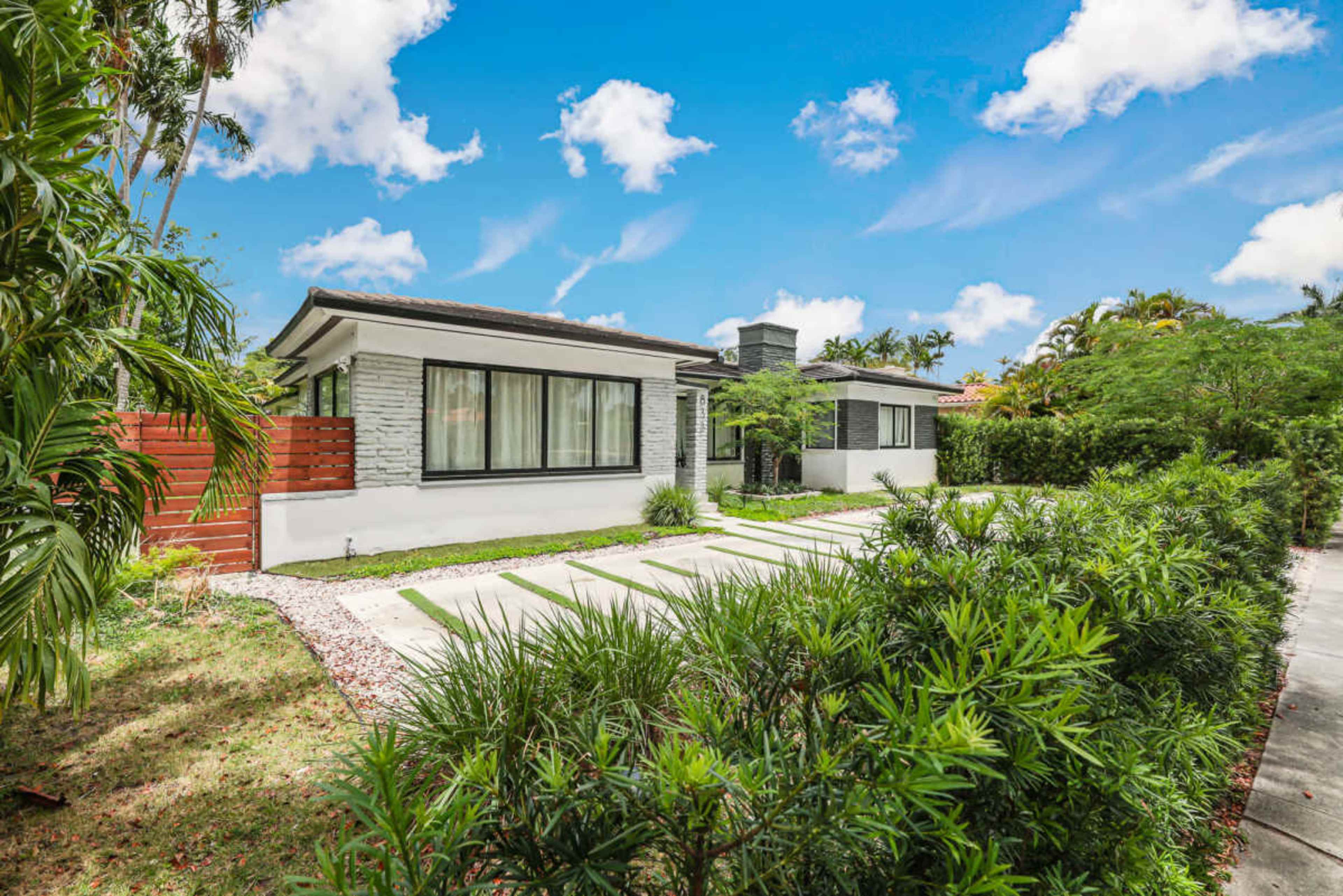 A modern single-story house with a stone facade and large windows is set amidst lush greenery and a neatly paved driveway.