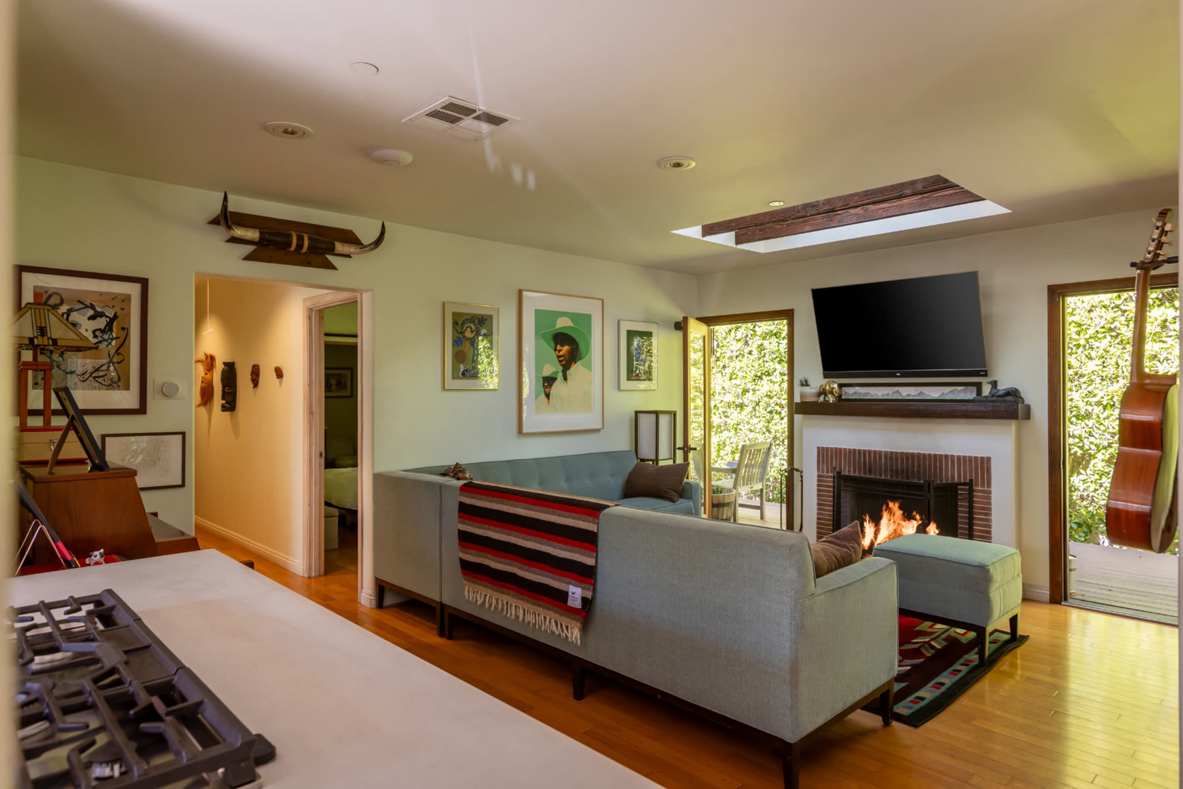 The image shows a living room with a modern couch, a fireplace, a mounted television, and a view of greenery through large windows.