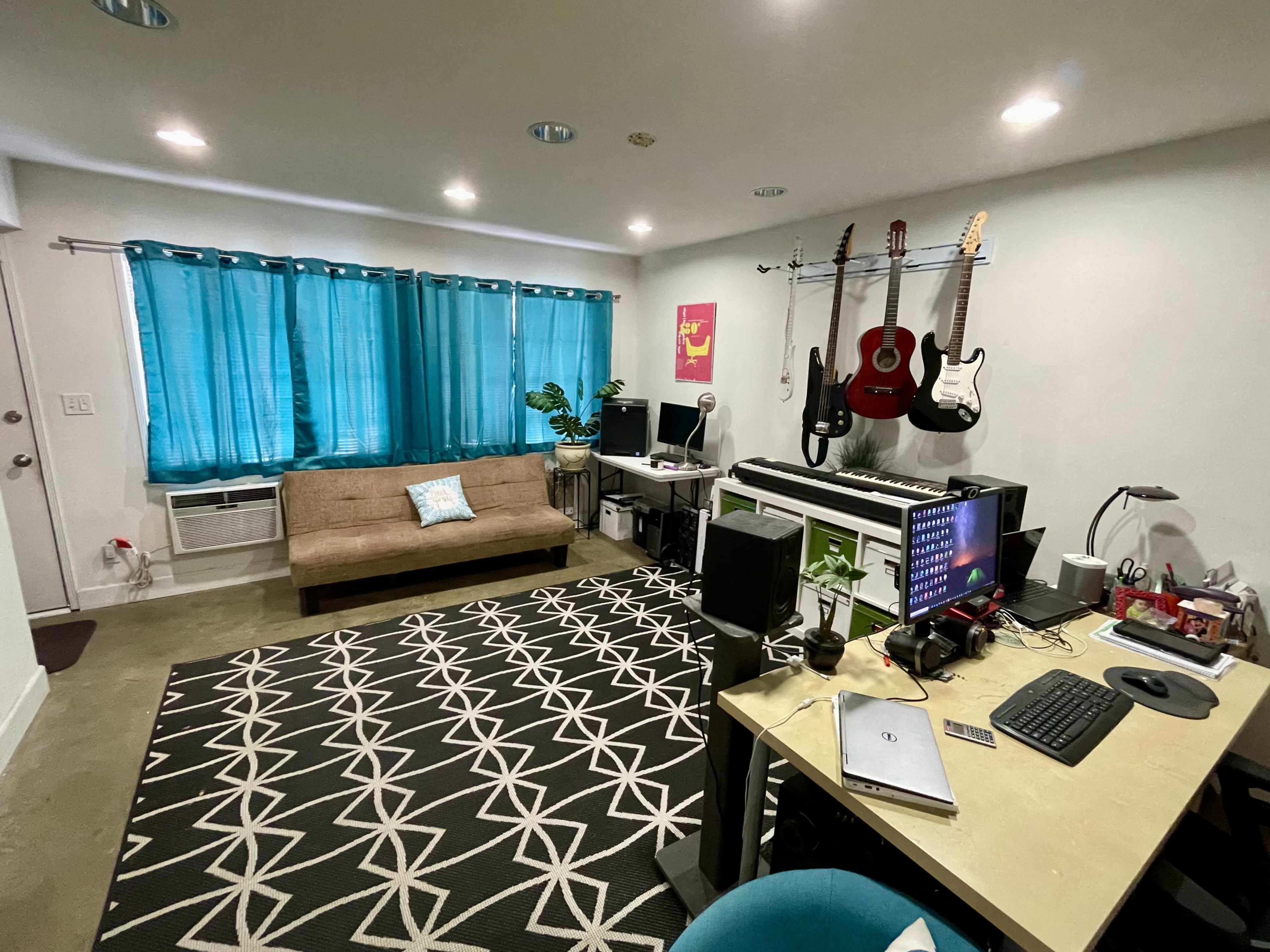 The image shows a small, modern living space with a couch, a desk with a computer, musical instruments on a rack, and blue curtains covering the windows.