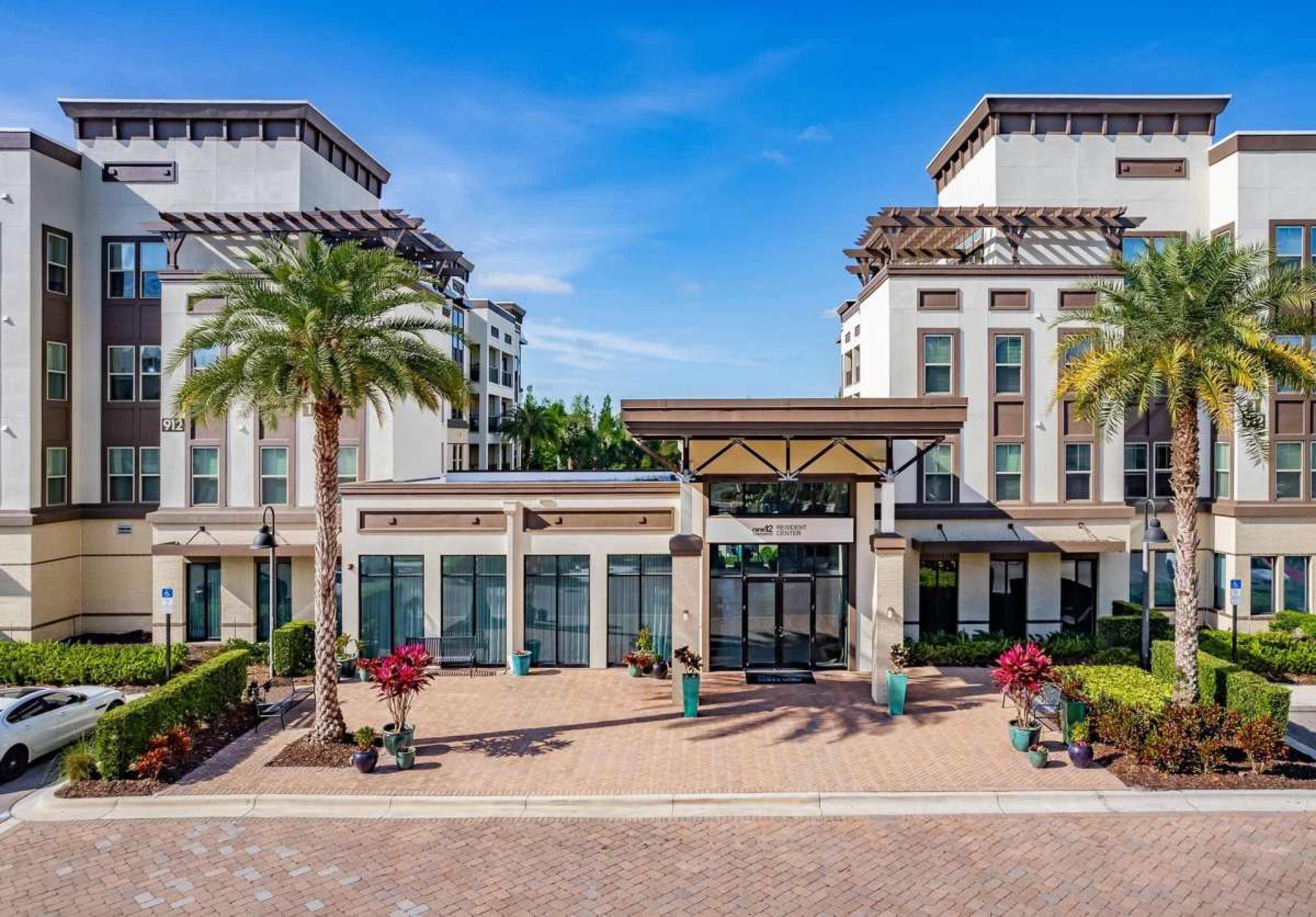 The image shows a modern apartment complex entrance flanked by palm trees and colorful plants, with a brick-paved pathway leading to the glass doors.