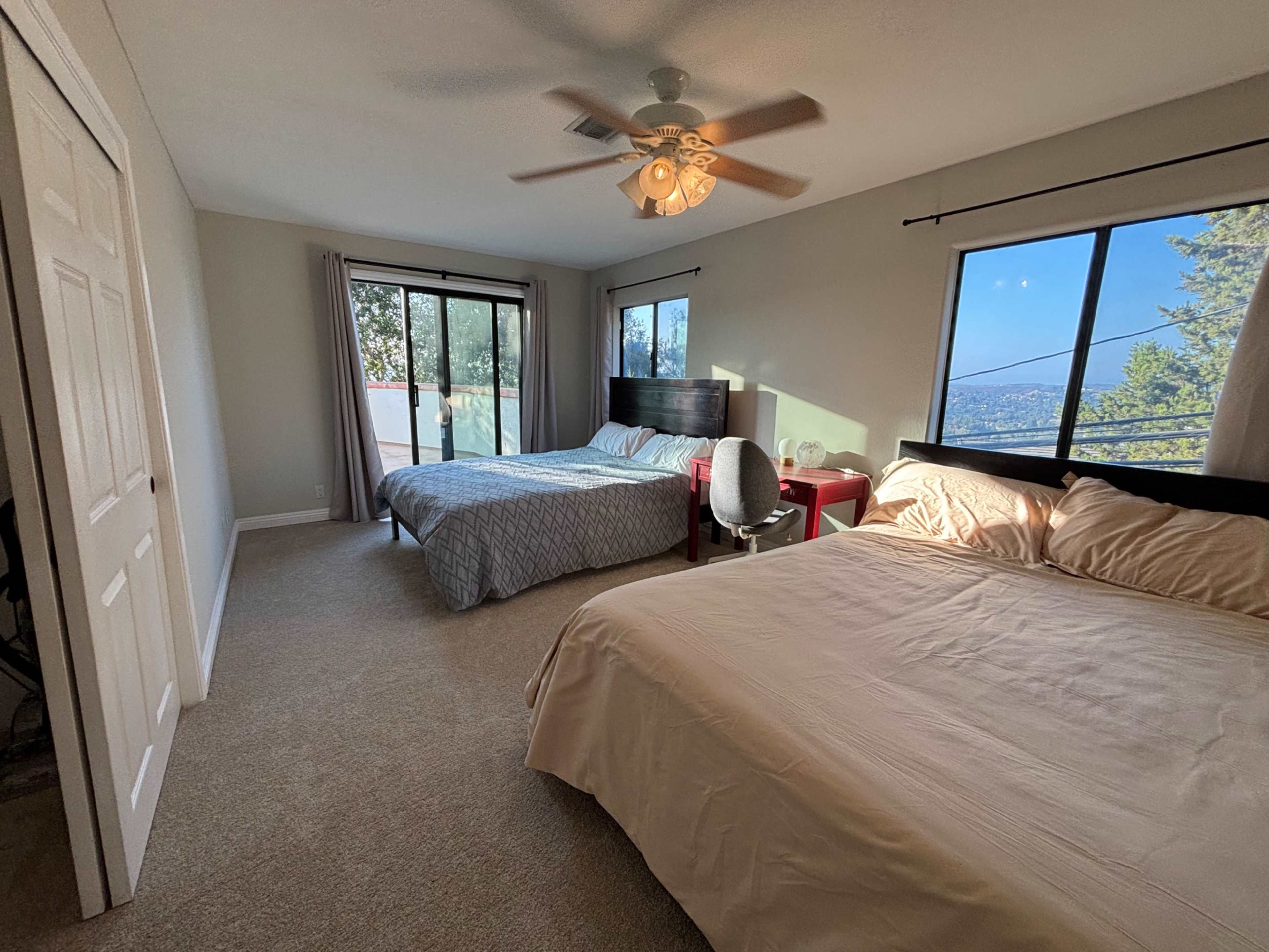 The image shows a spacious bedroom featuring two beds, a ceiling fan, and large windows with a view of trees and a distant landscape.