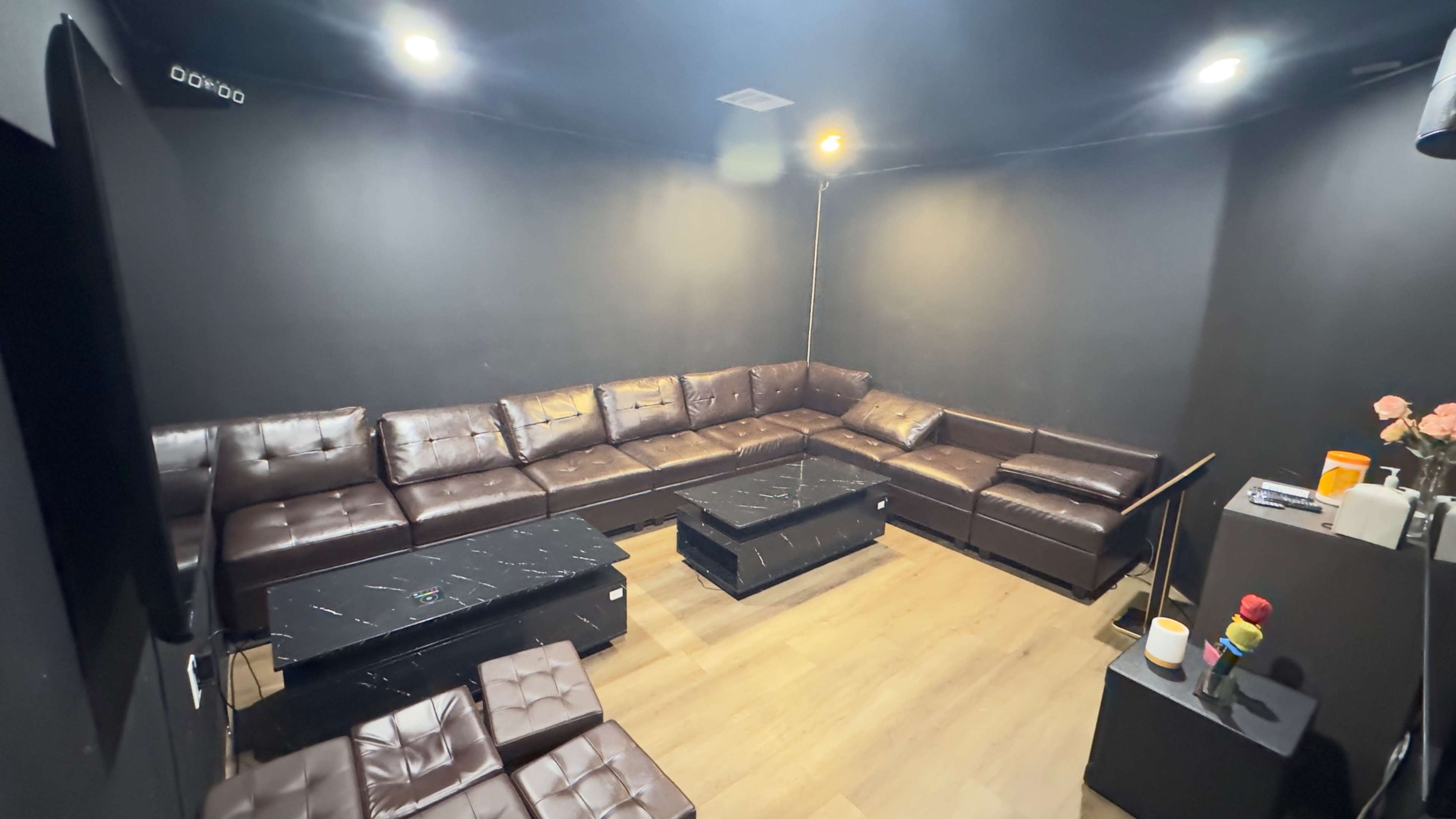The image shows a small, dimly lit lounge area with dark walls, a large brown sectional sofa, and two black coffee tables.