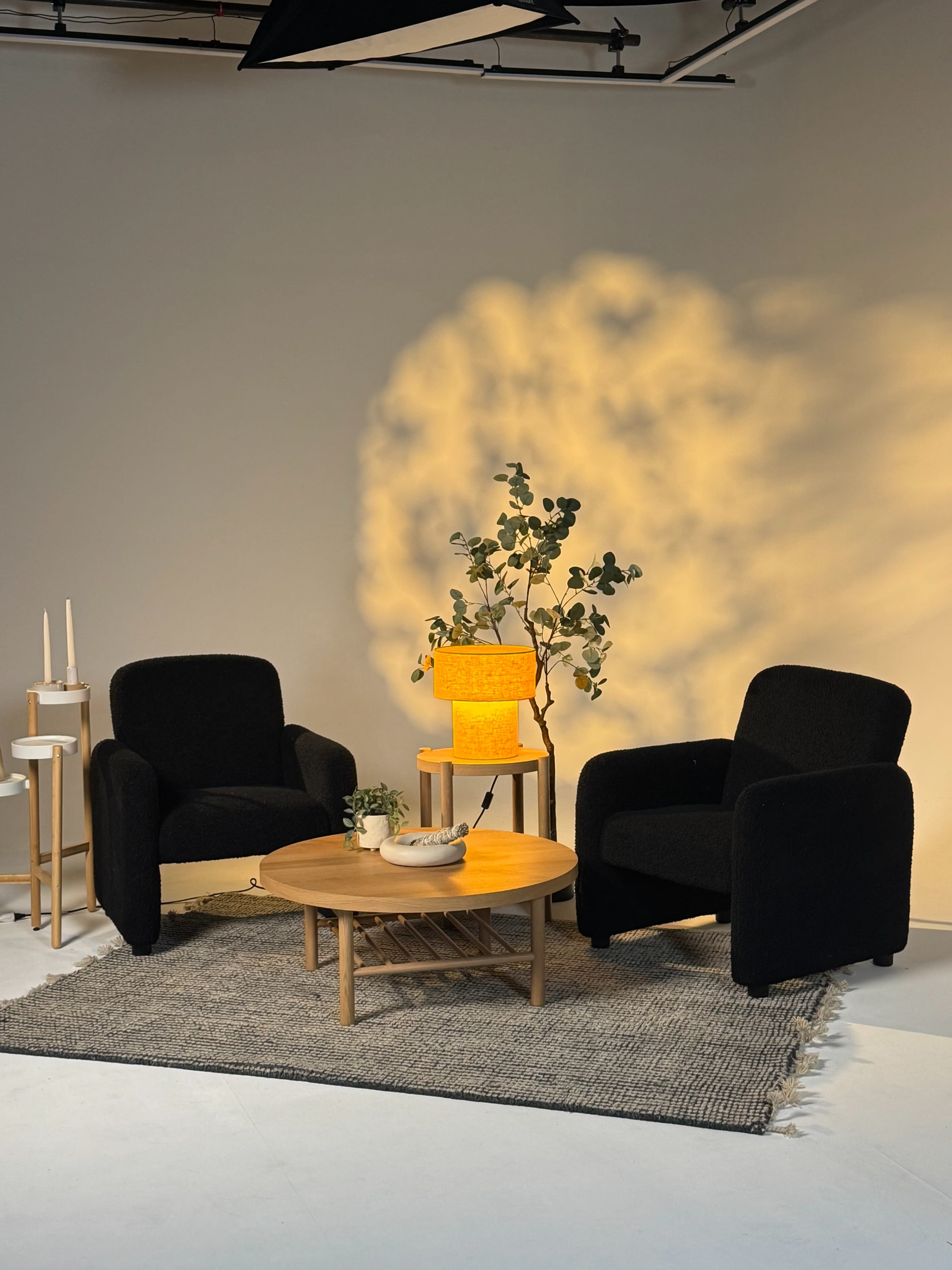 The image features two black armchairs on a textured rug, a round wooden coffee table, a yellow lamp, and a small table with candles beside a potted plant in a softly lit indoor setting.