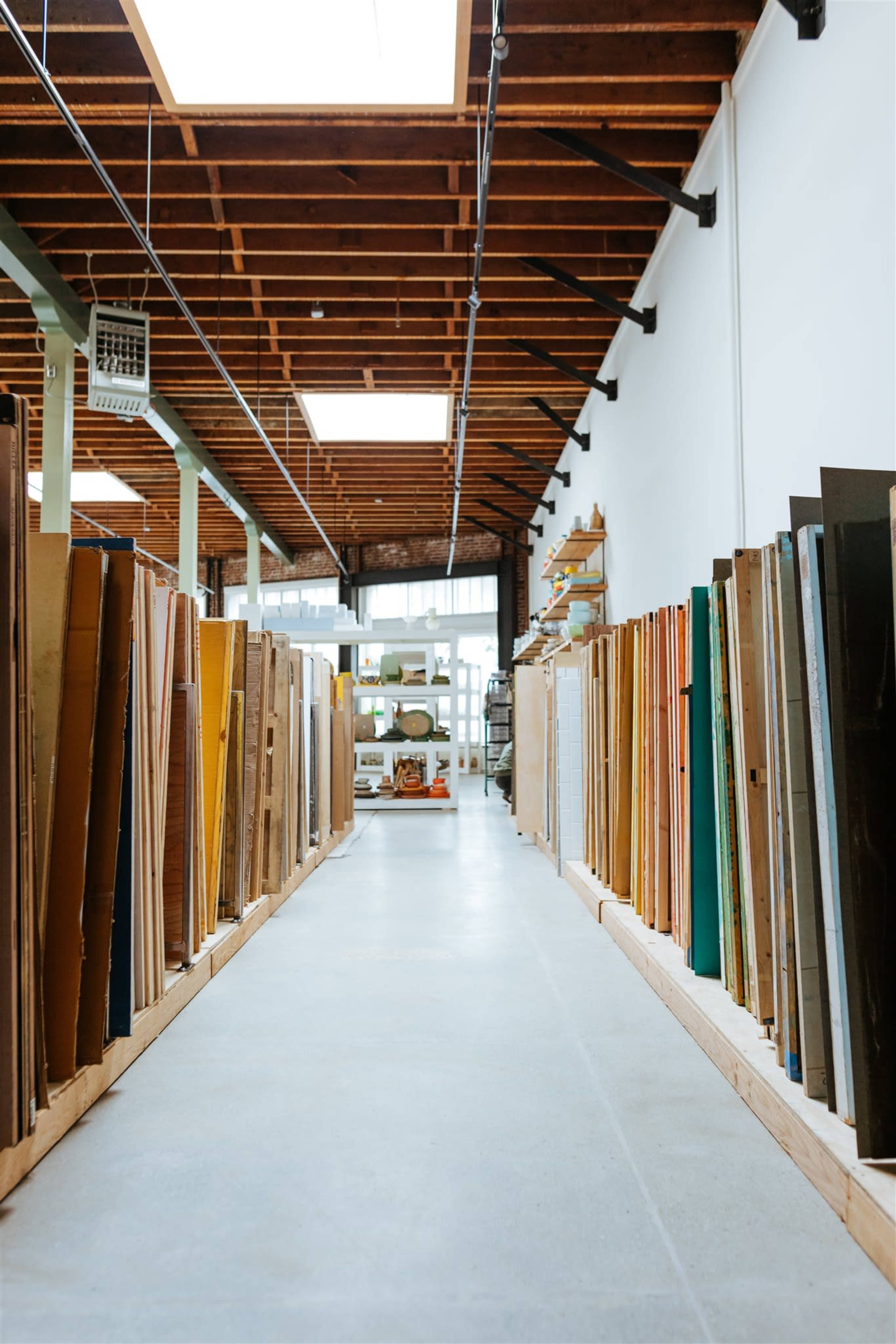 A wide aisle between tall stacks of various boards in a spacious workshop with a wooden ceiling.