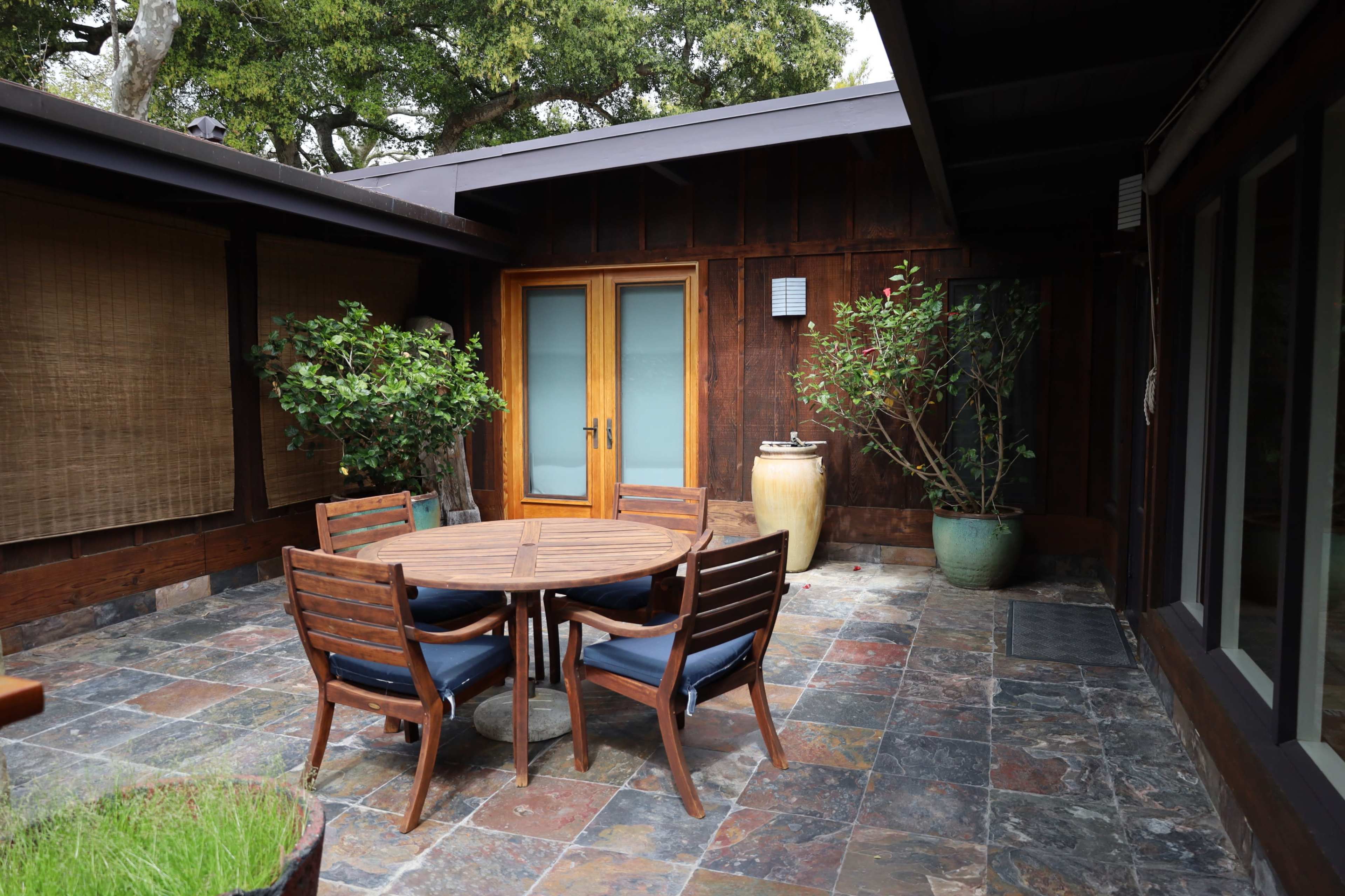 The image shows a patio area with a round wooden table surrounded by six chairs, flanked by potted plants and a large ceramic vase.