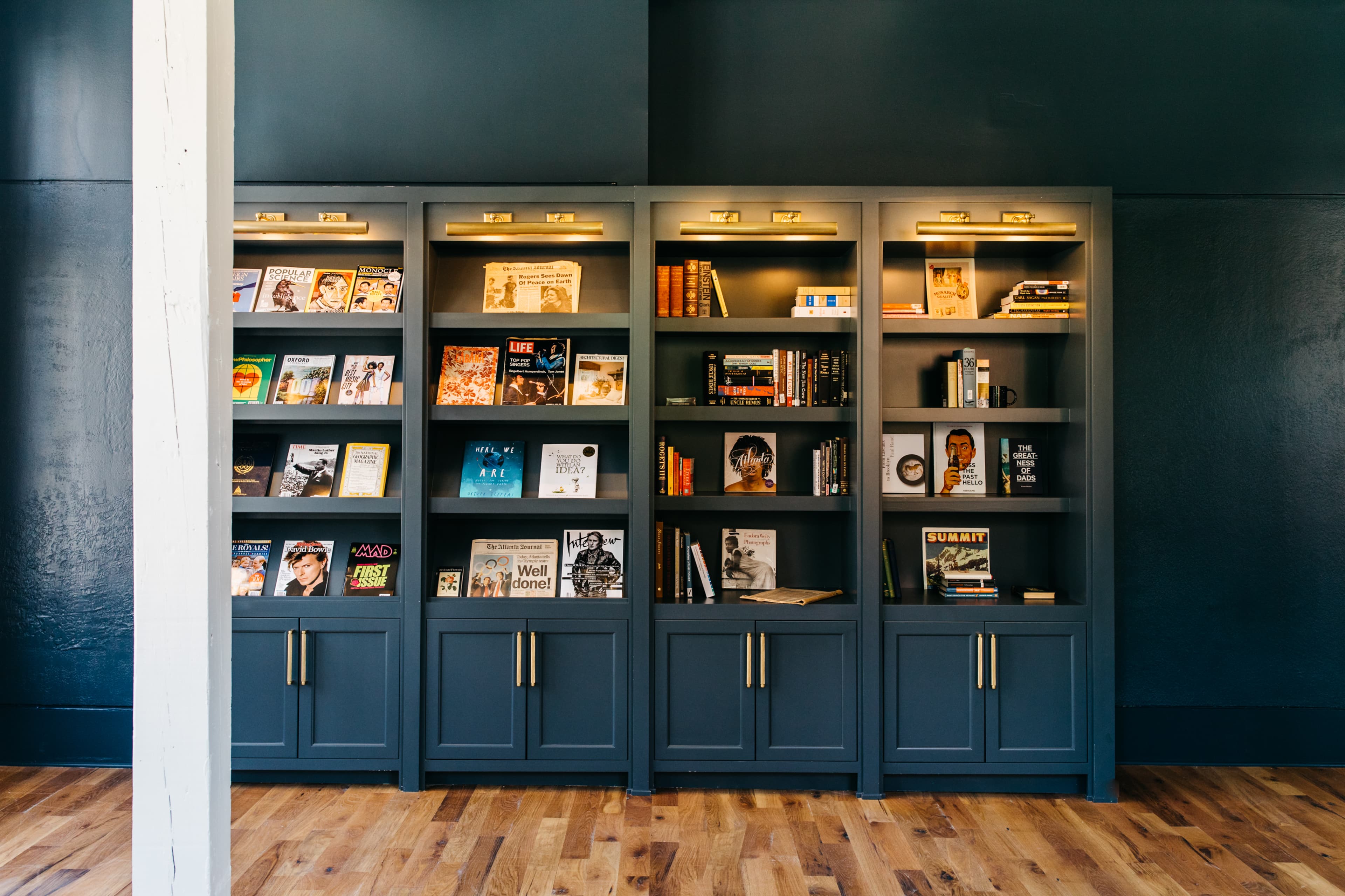 A tall, dark gray bookshelf holds various books and magazines against a dark wall in a room with wooden flooring.