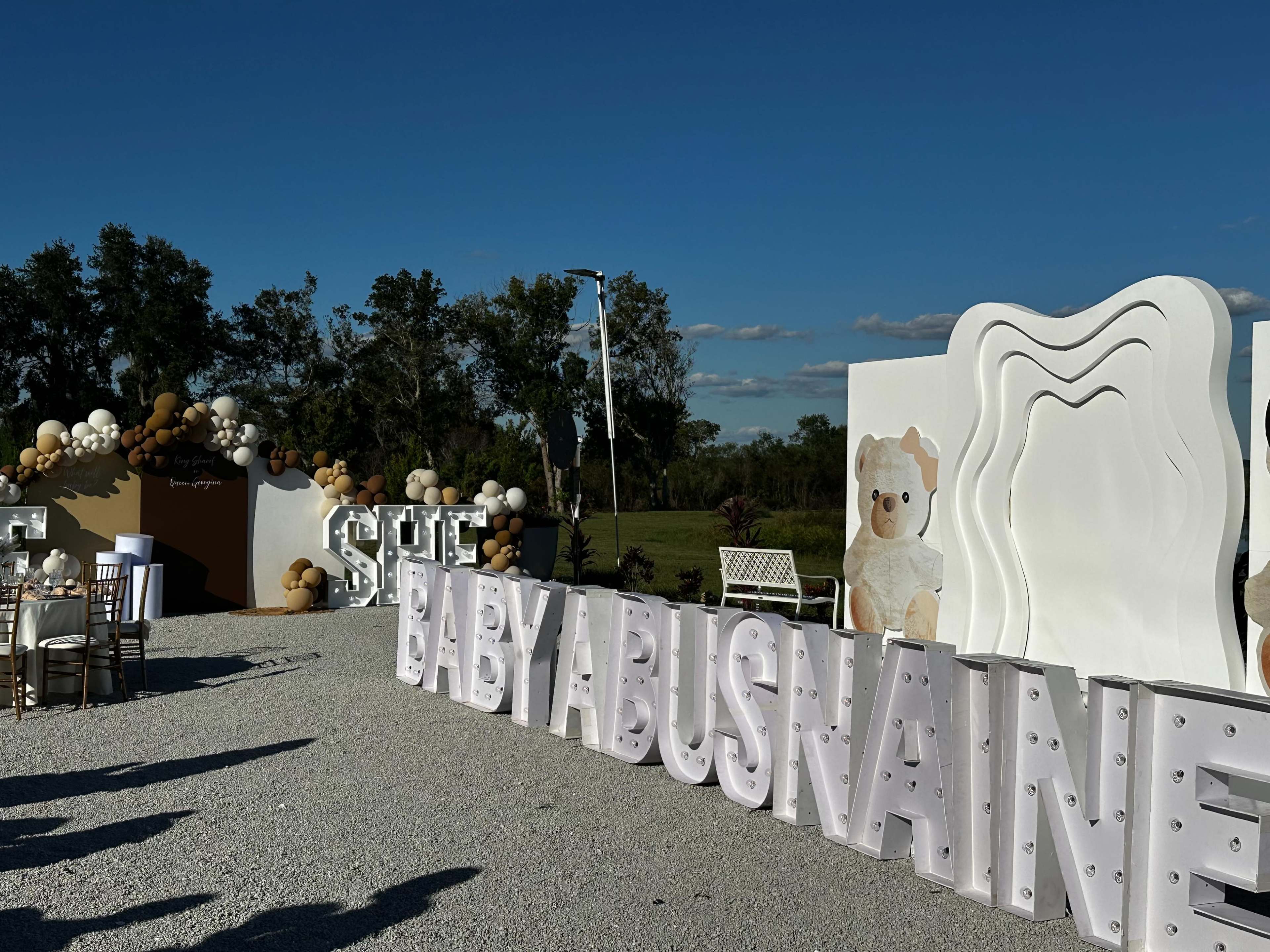 The scene features a decorated outdoor area with large illuminated letters spelling "BABY ABUSNAINE," surrounded by balloon arrangements and a backdrop with a teddy bear graphic.