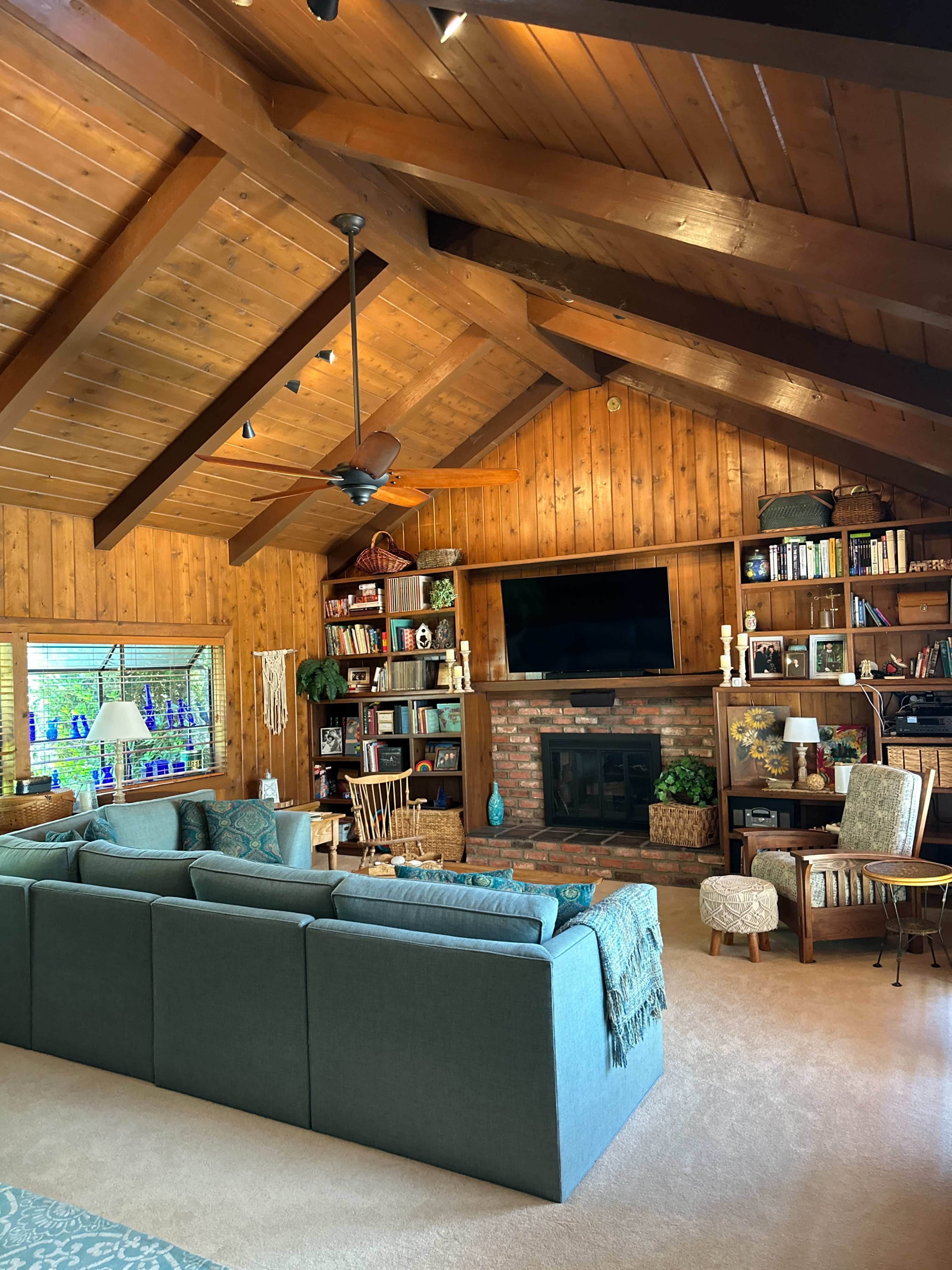 A cozy living room features a large sectional sofa, a brick fireplace, wooden beams, and built-in bookshelves filled with books and decor.