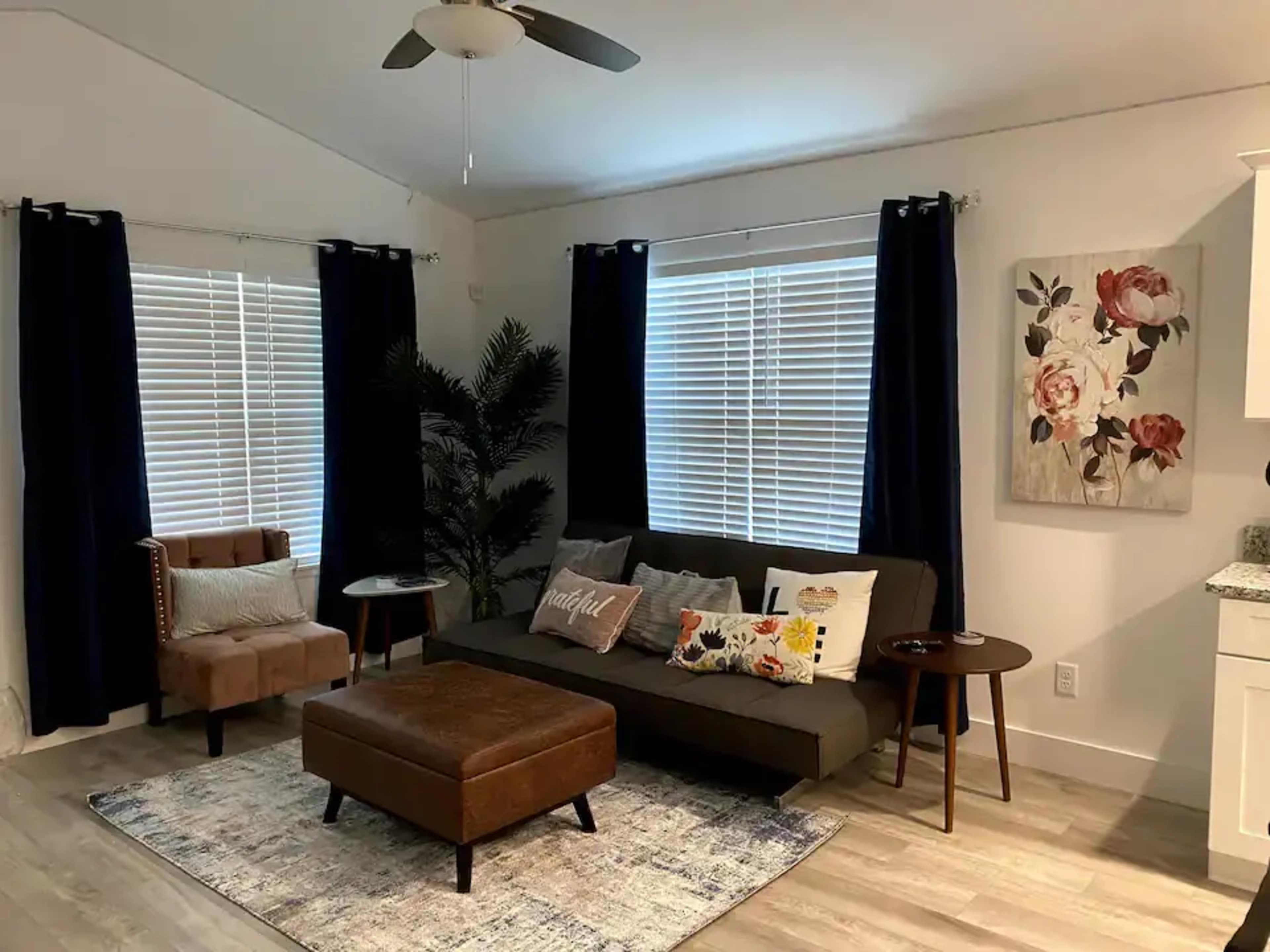 A modern living room with a couch, armchair, and coffee table, featuring large windows with white blinds and dark curtains.