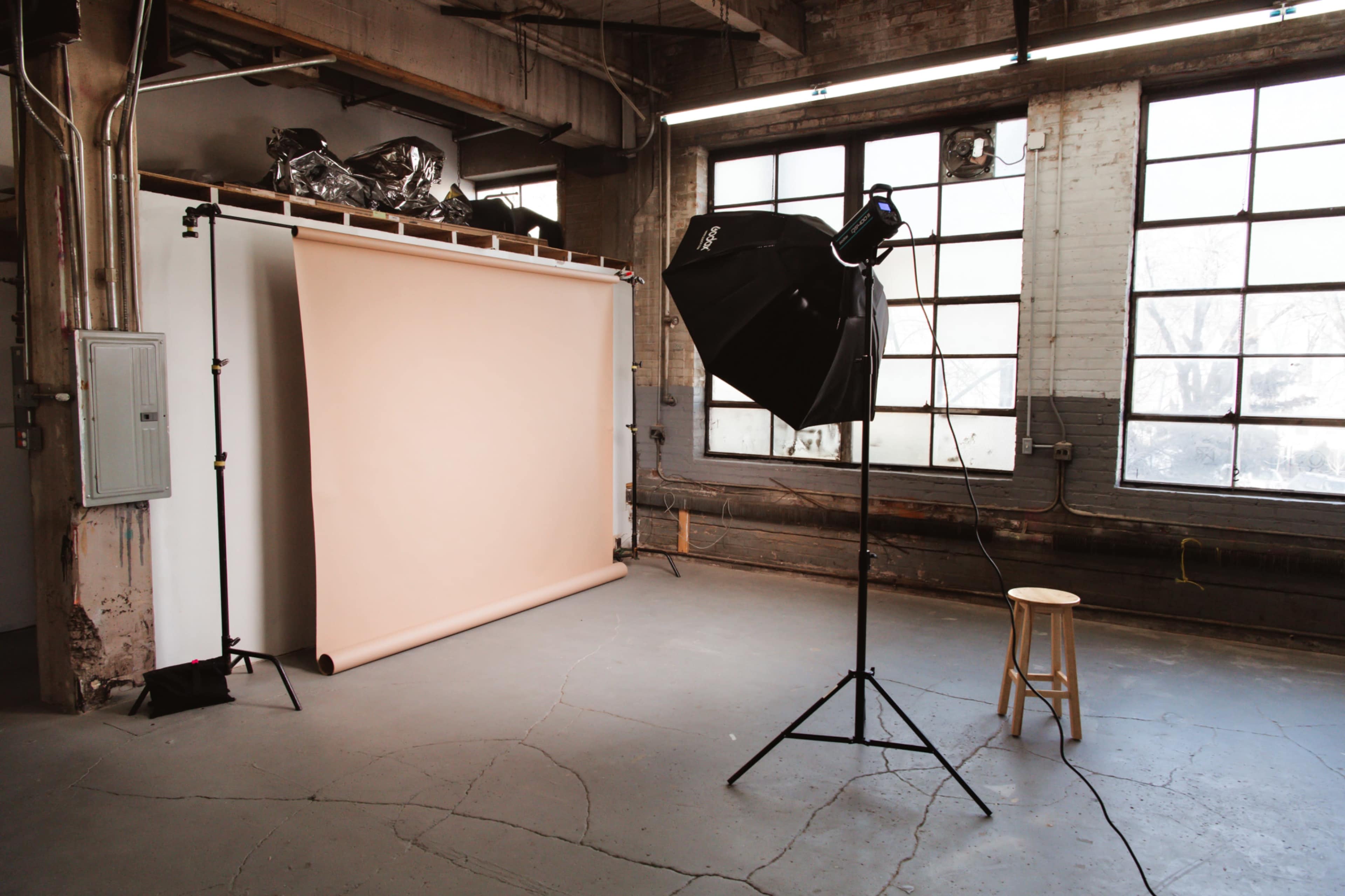 A photography studio featuring a light stand, a beige backdrop, and a stool against large windows.