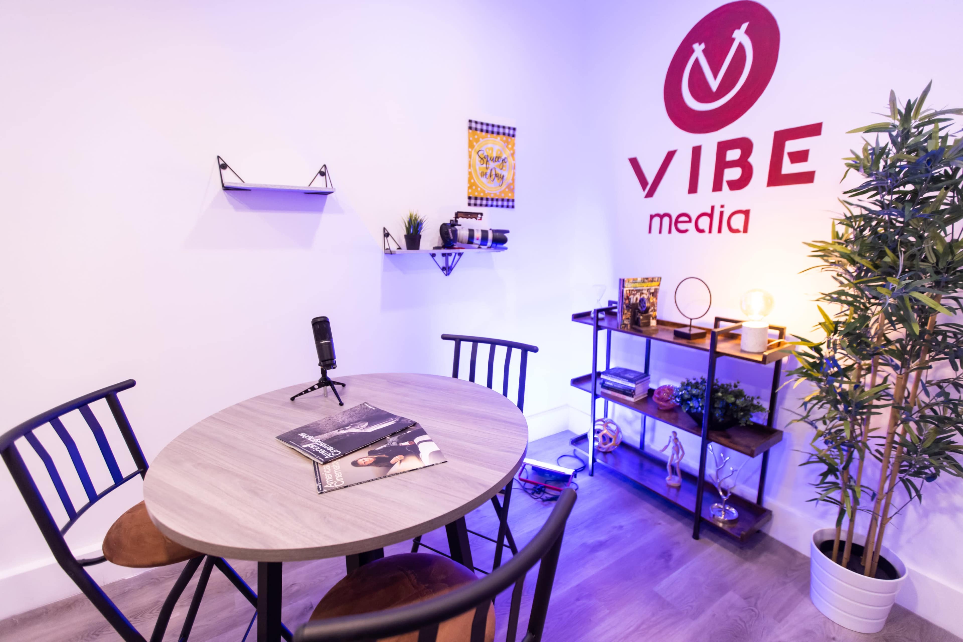 The image shows a small modern media office with a round table, chairs, a microphone, and shelves displaying various items against a white wall decorated with the "VIBE media" logo.