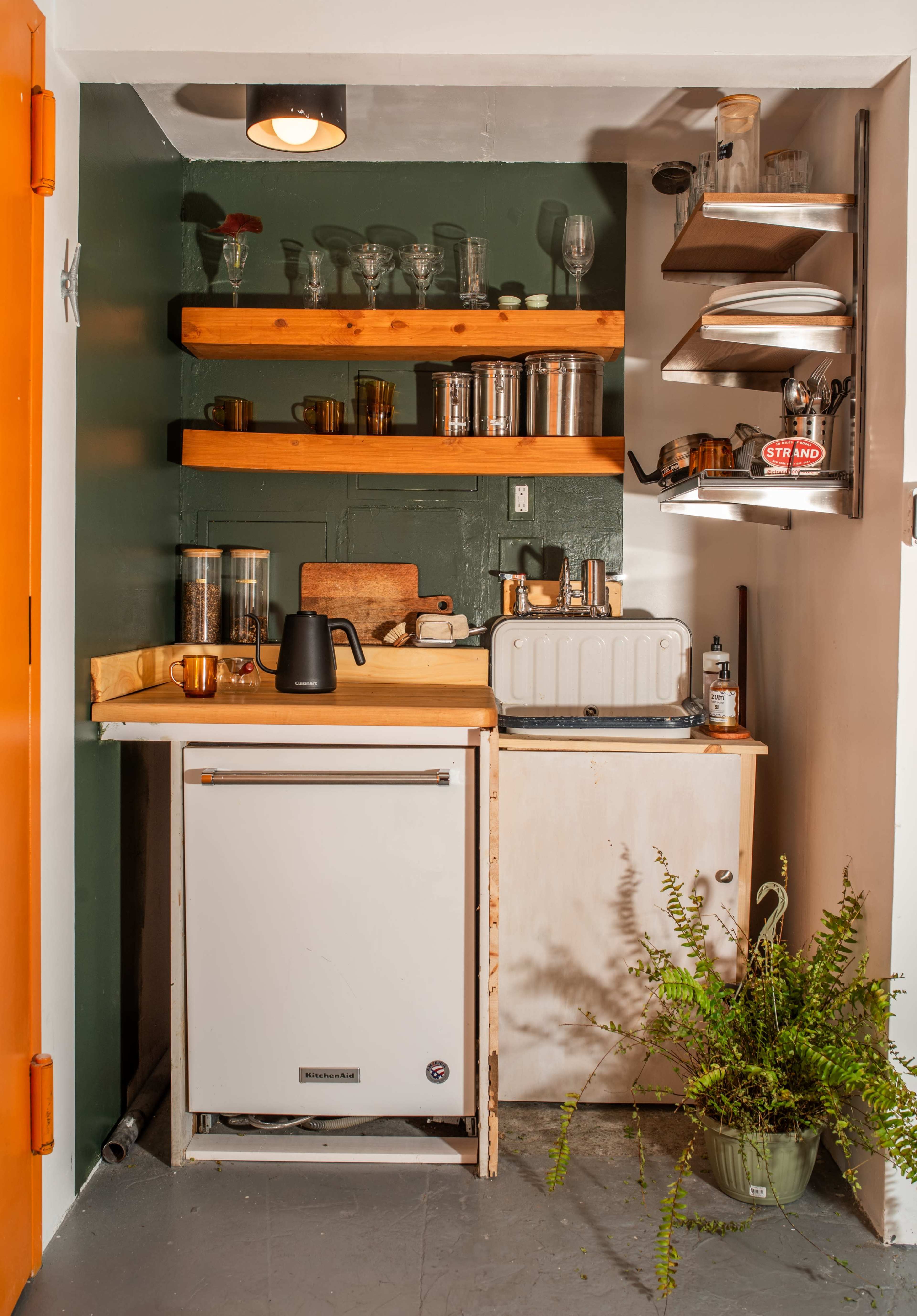 The image shows a compact kitchen area featuring a wooden countertop with kitchen supplies, glassware on floating shelves, and an under-counter refrigerator beside a small potted plant.