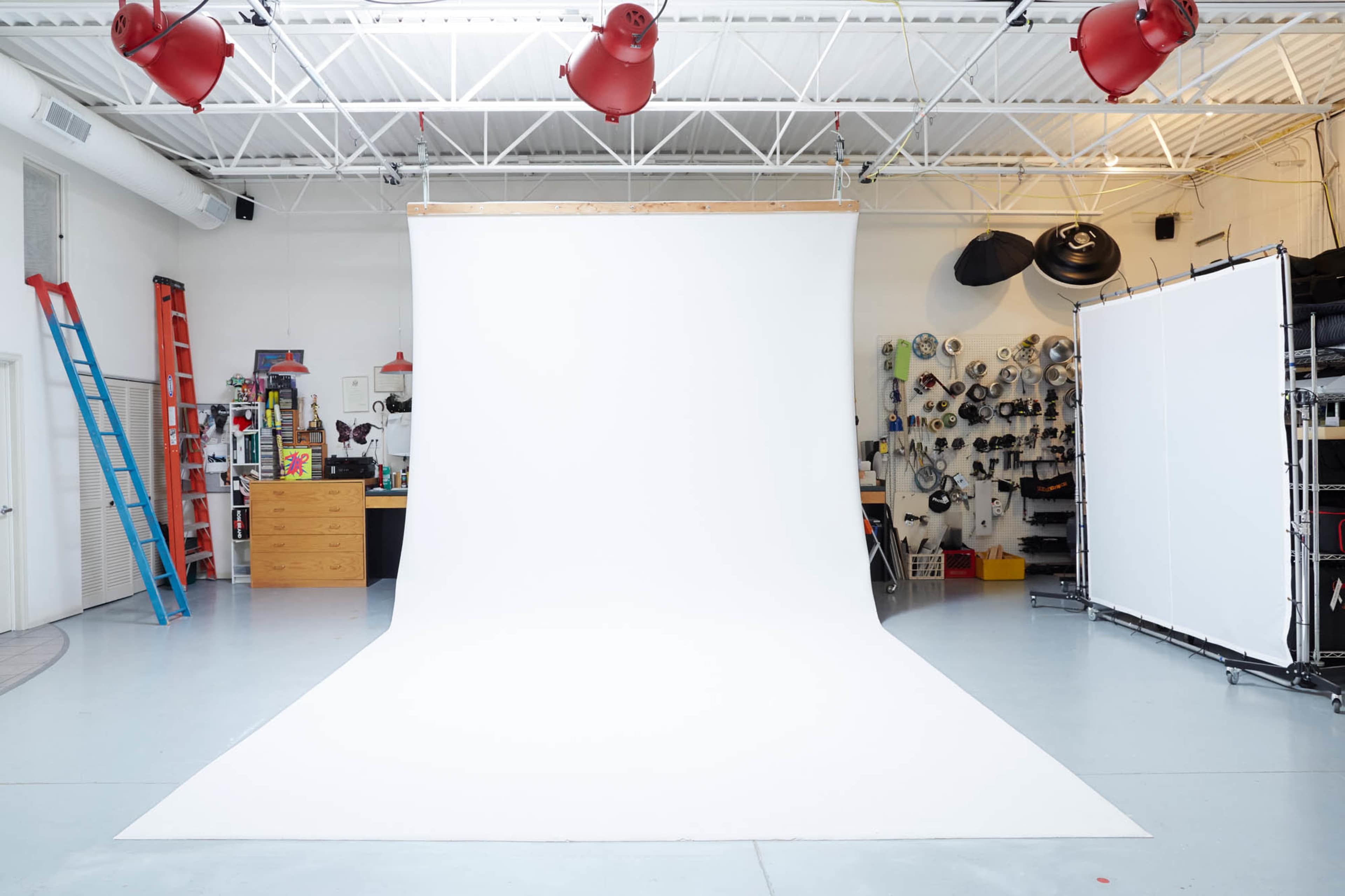 A spacious photography studio features a white backdrop, a ladder, and various equipment along the walls.