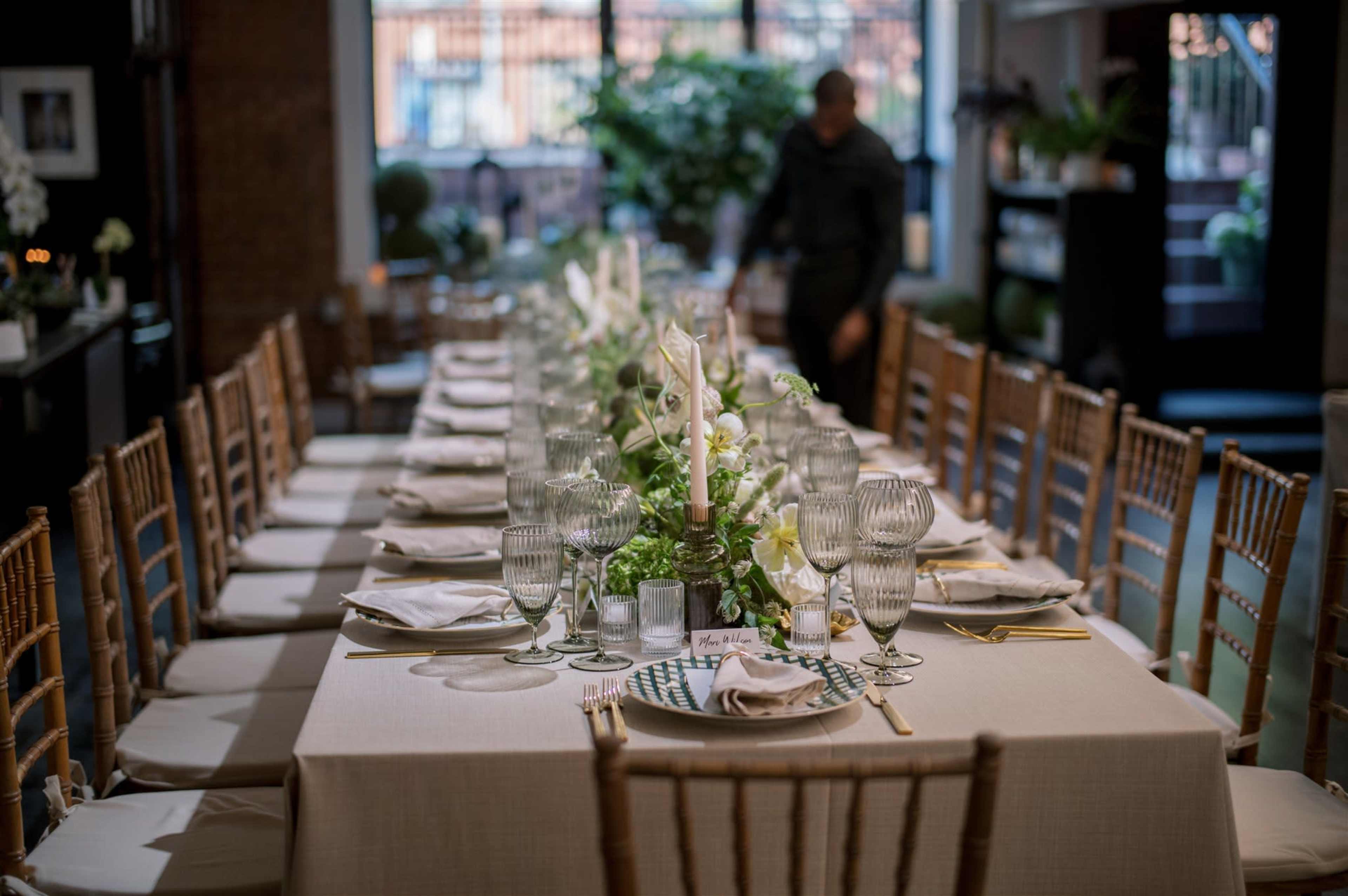 A long dining table is set with fine china, crystal glassware, and floral centerpieces, while a person adjusts items in the background.