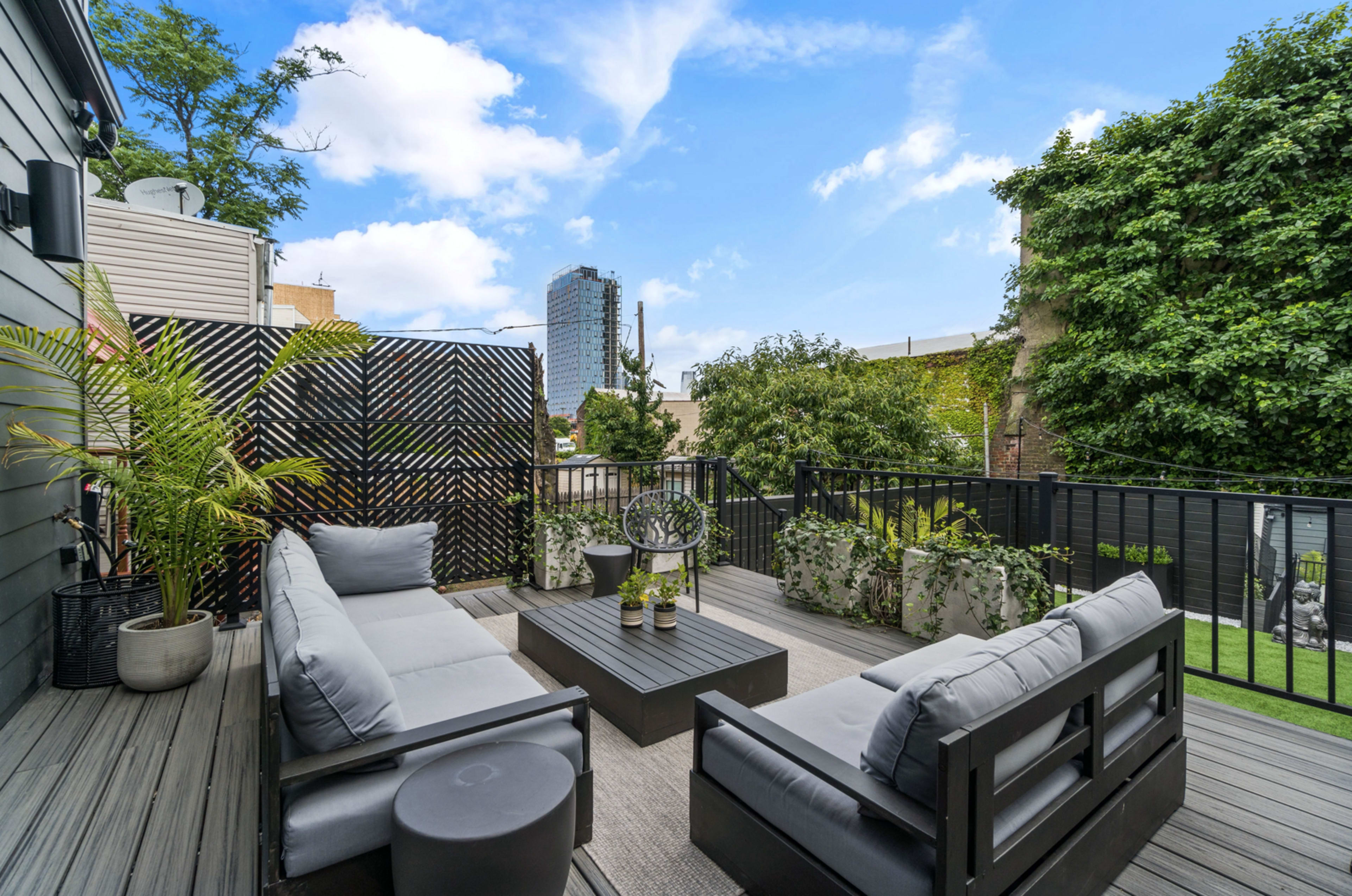 A balcony with gray outdoor seating, a coffee table, and various plants, overlooking a cityscape with tall buildings in the background.