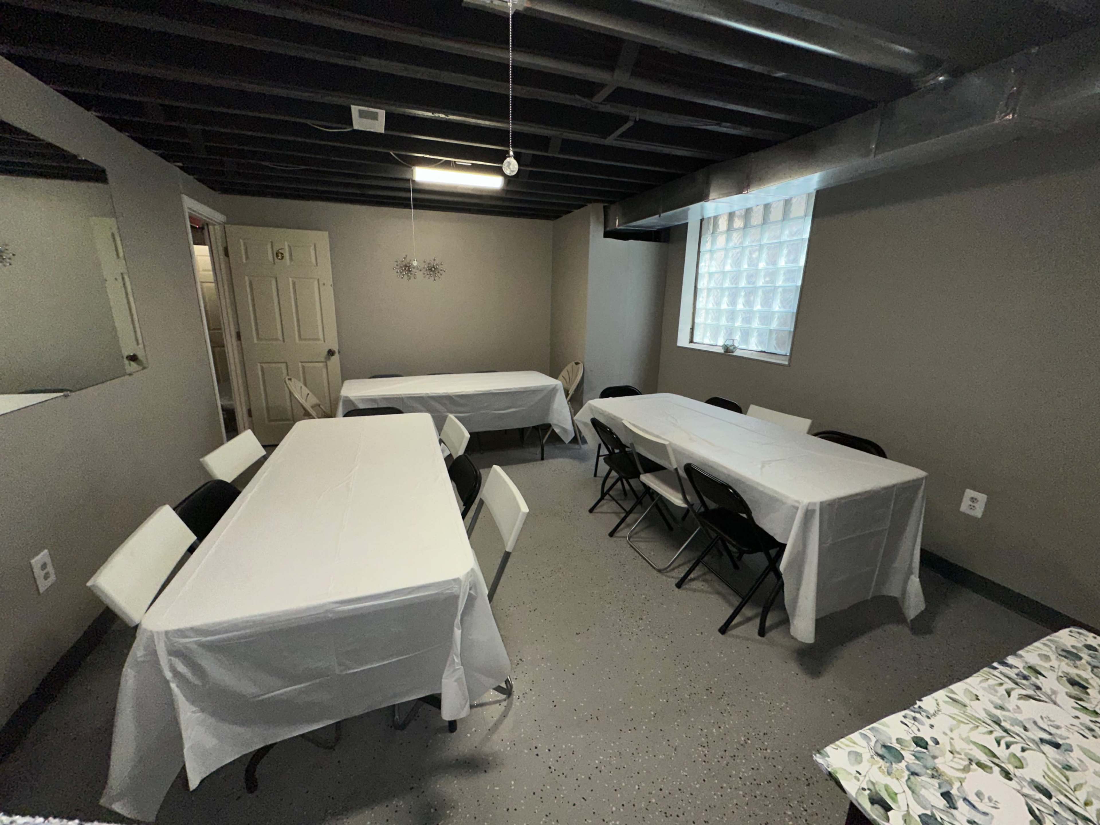 The image shows a basement room with several long tables covered in white tablecloths and chairs arranged around them, along with a mirror and a window featuring a glass block design.