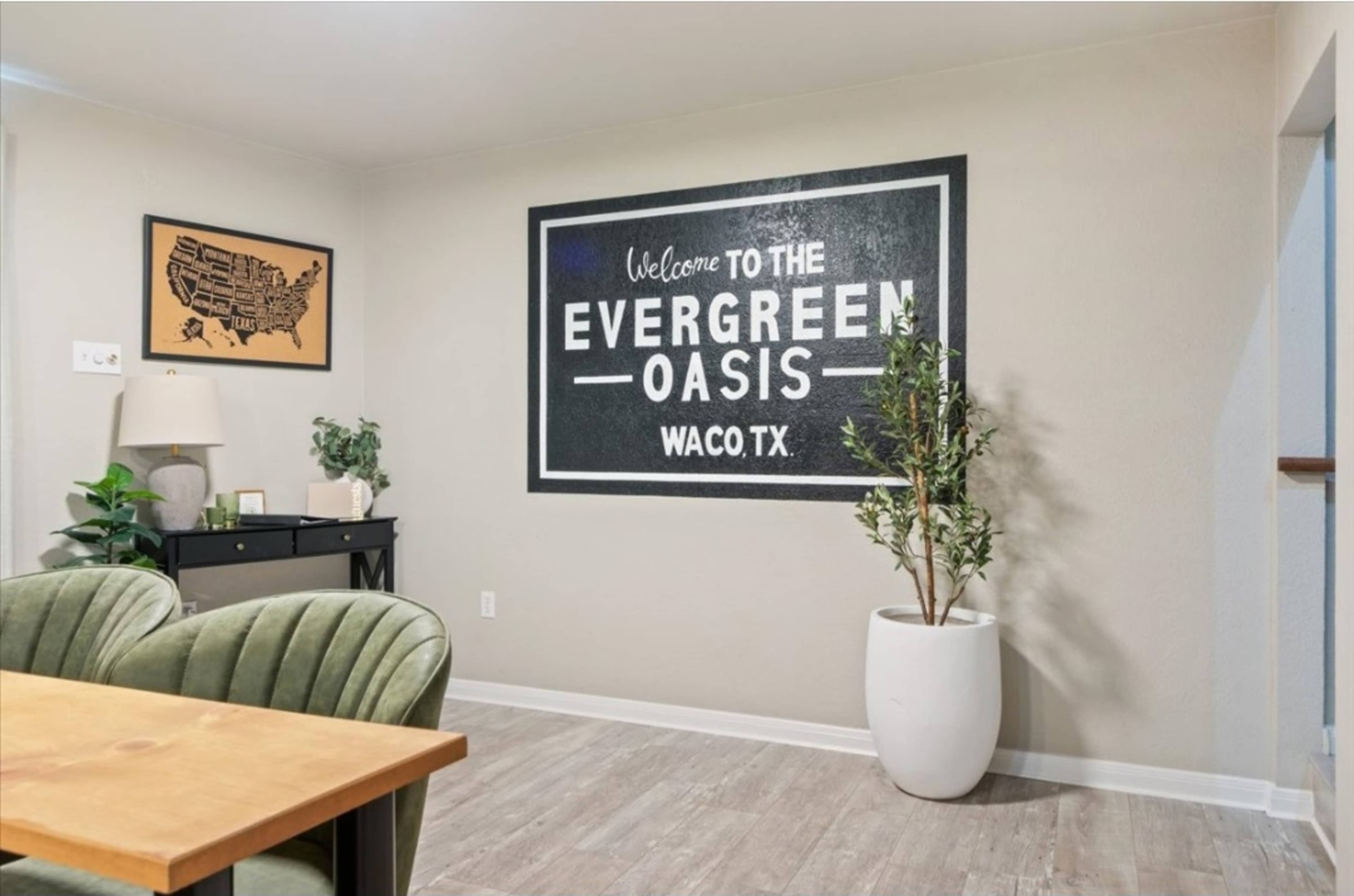 A modern room featuring a wall with a large sign that reads "Welcome to the Evergreen Oasis Waco, TX," alongside a dining table and a decorative plant in a white pot.