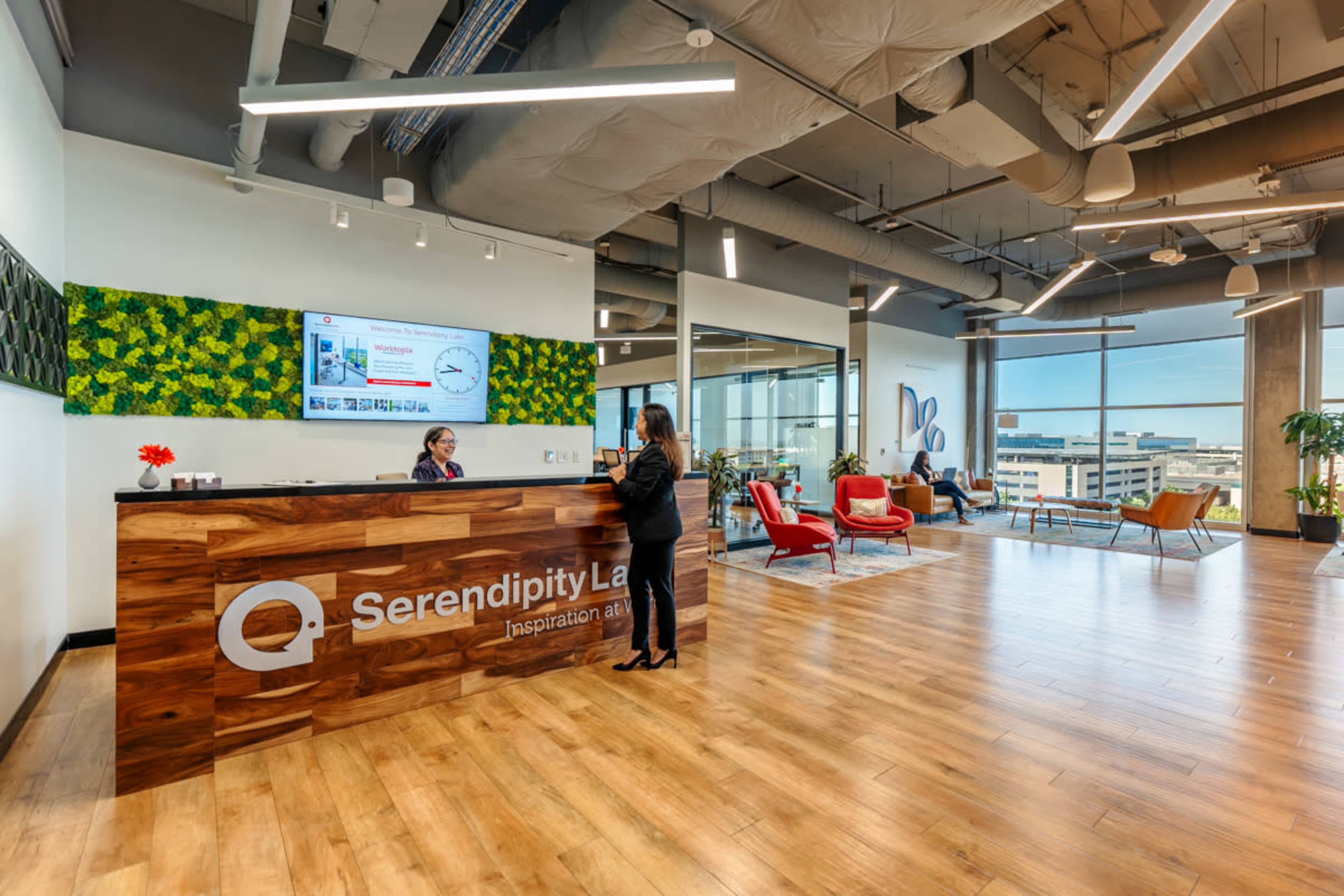 A modern reception area features a wooden front desk with the name "Serendipity Labs," surrounded by stylish seating and large windows offering a view of the city.