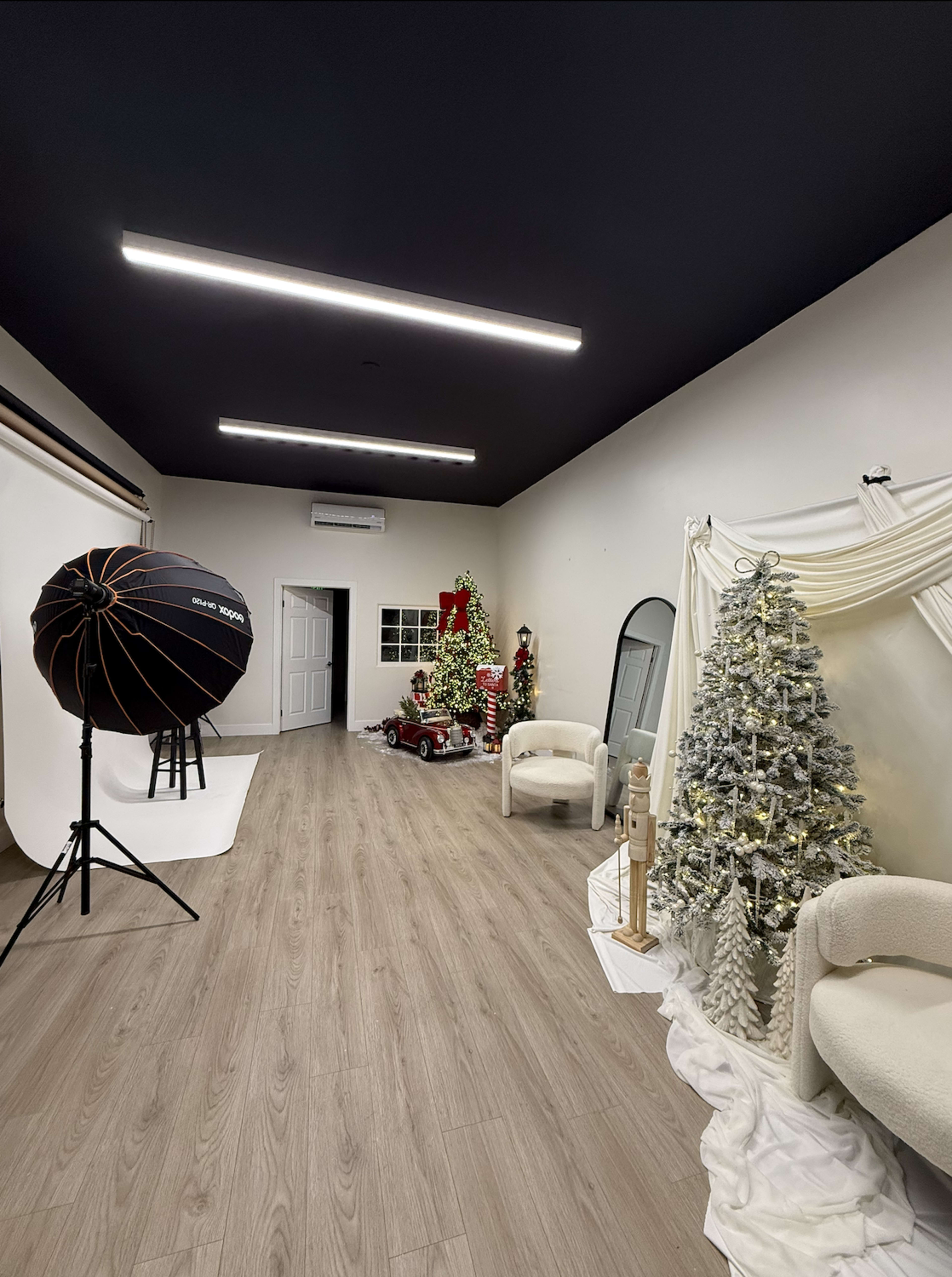 The image shows a photography studio decorated for the holidays, featuring a Christmas tree, a vintage toy car, and various props arranged beside a backdrop stand.