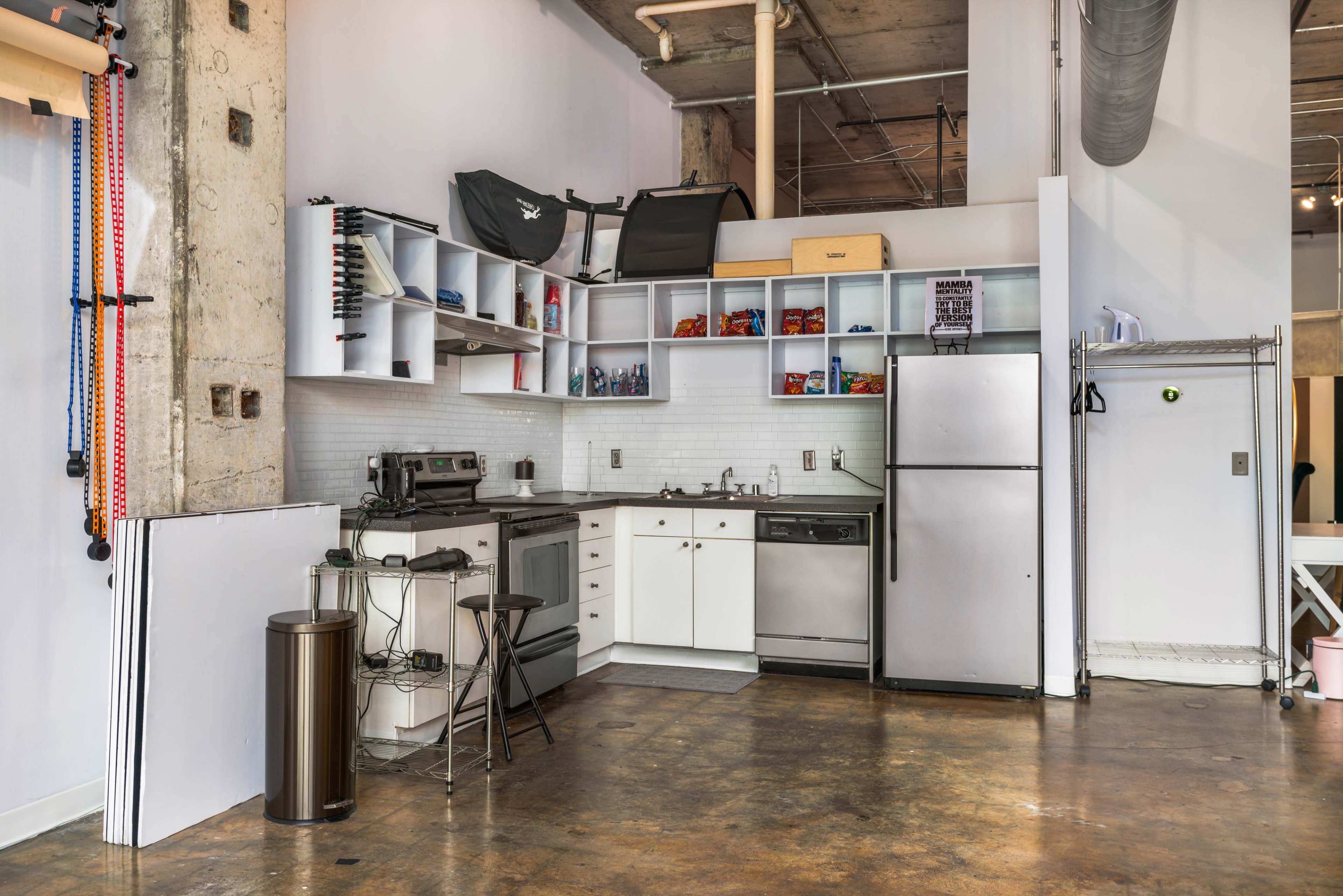A compact kitchen area featuring a stove, oven, refrigerator, and shelving, with industrial-style walls and flooring.