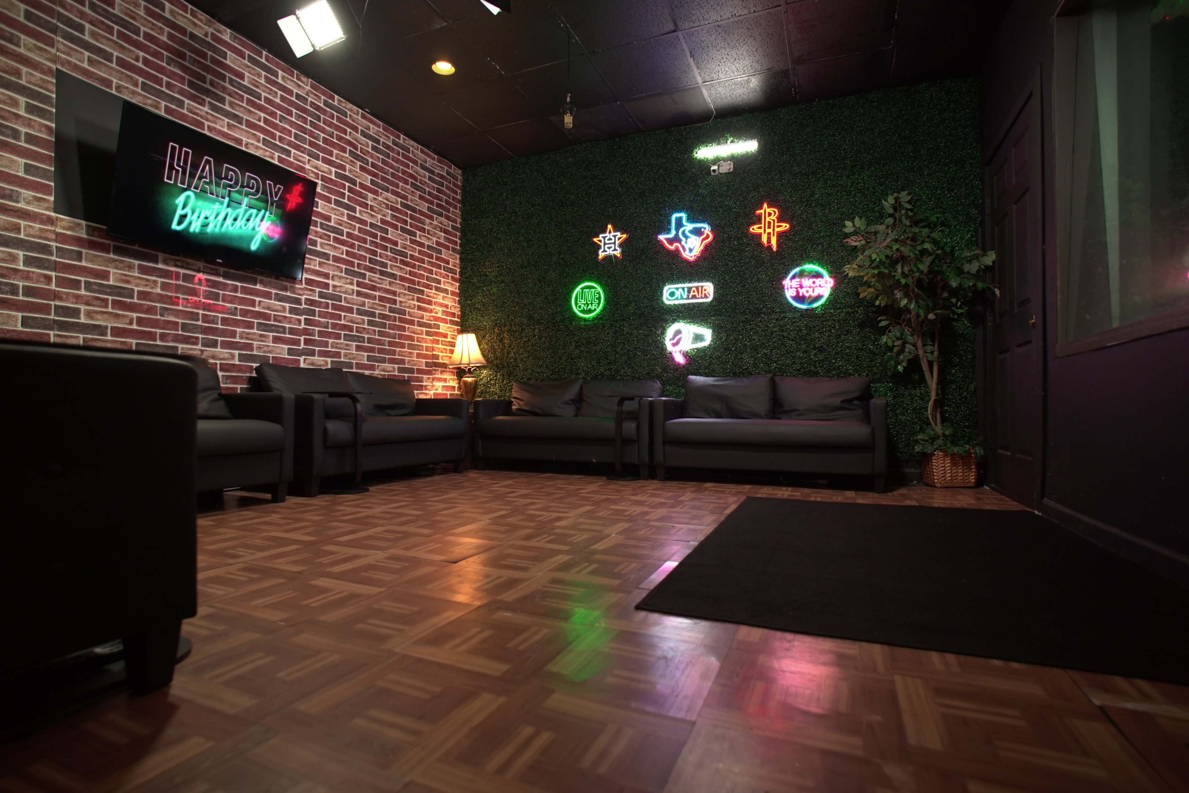 The image shows a cozy lounge area featuring brick walls, dark furniture, neon birthday-themed signs, and a wooden floor.