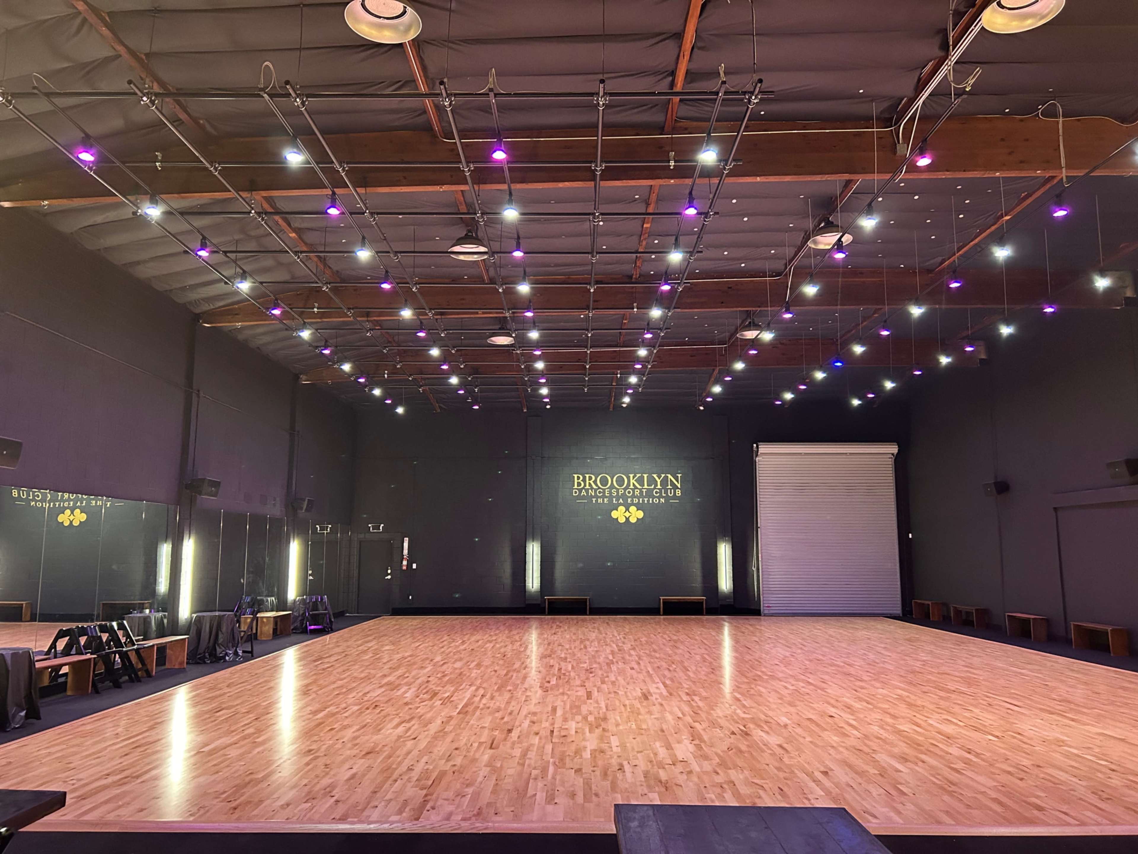 A large dance studio with a wooden floor, mirrored walls, and overhead lighting in purple shades.