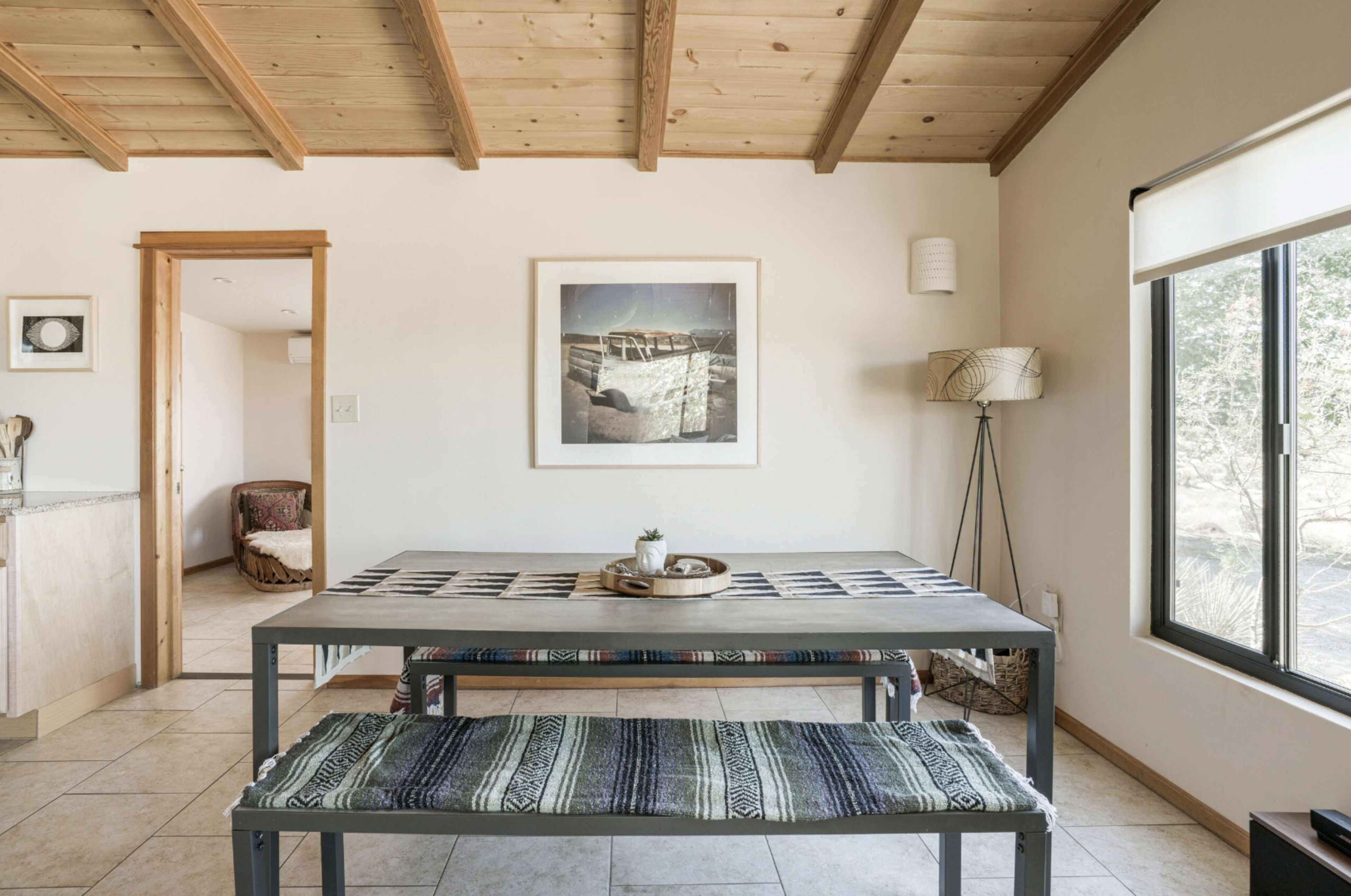 A dining area features a wooden table with patterned runners and benches, surrounded by neutral walls and a window with natural light.