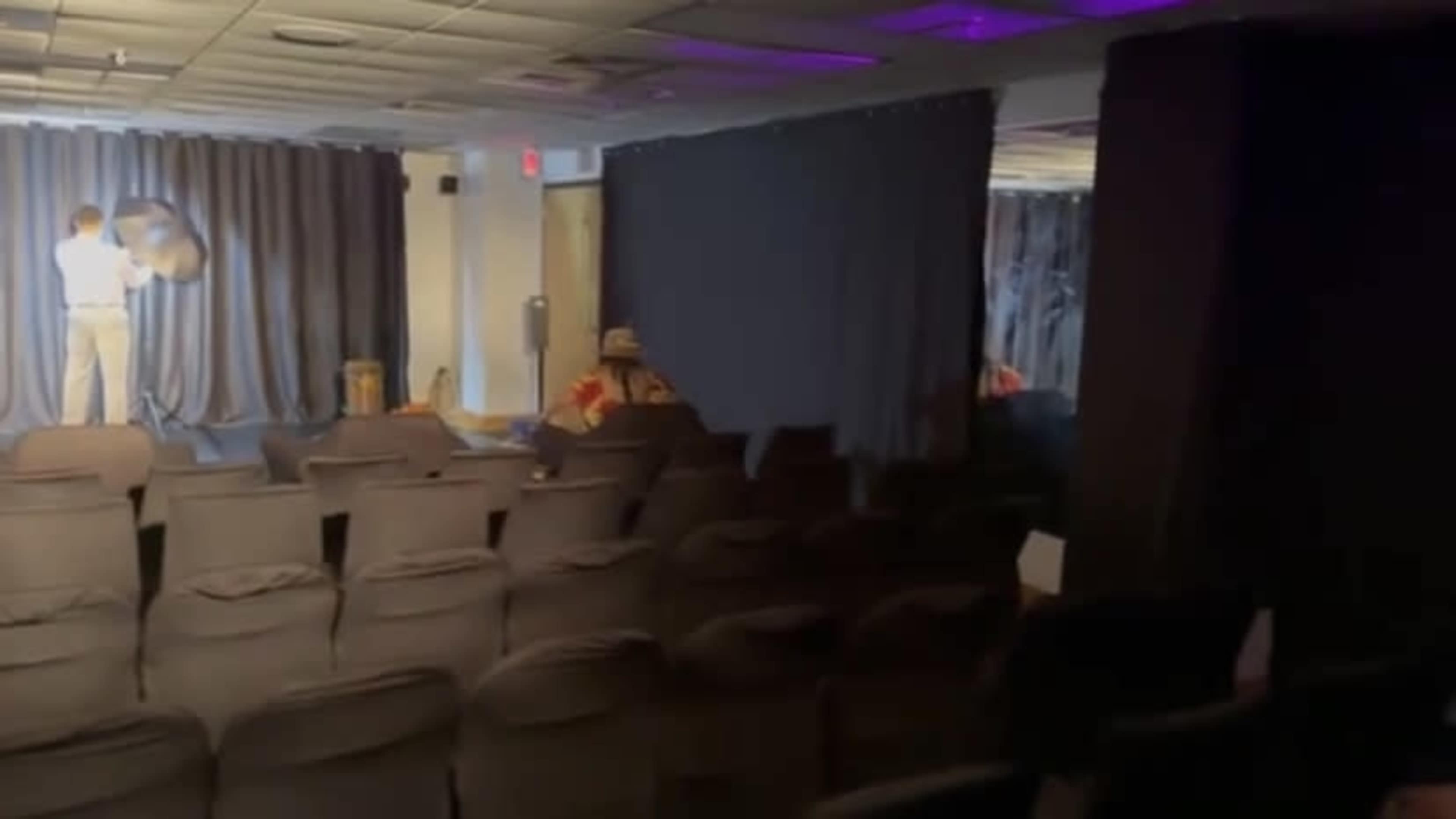 A dimly lit room features rows of black chairs facing a stage with curtains, where a person is adjusting a prop.