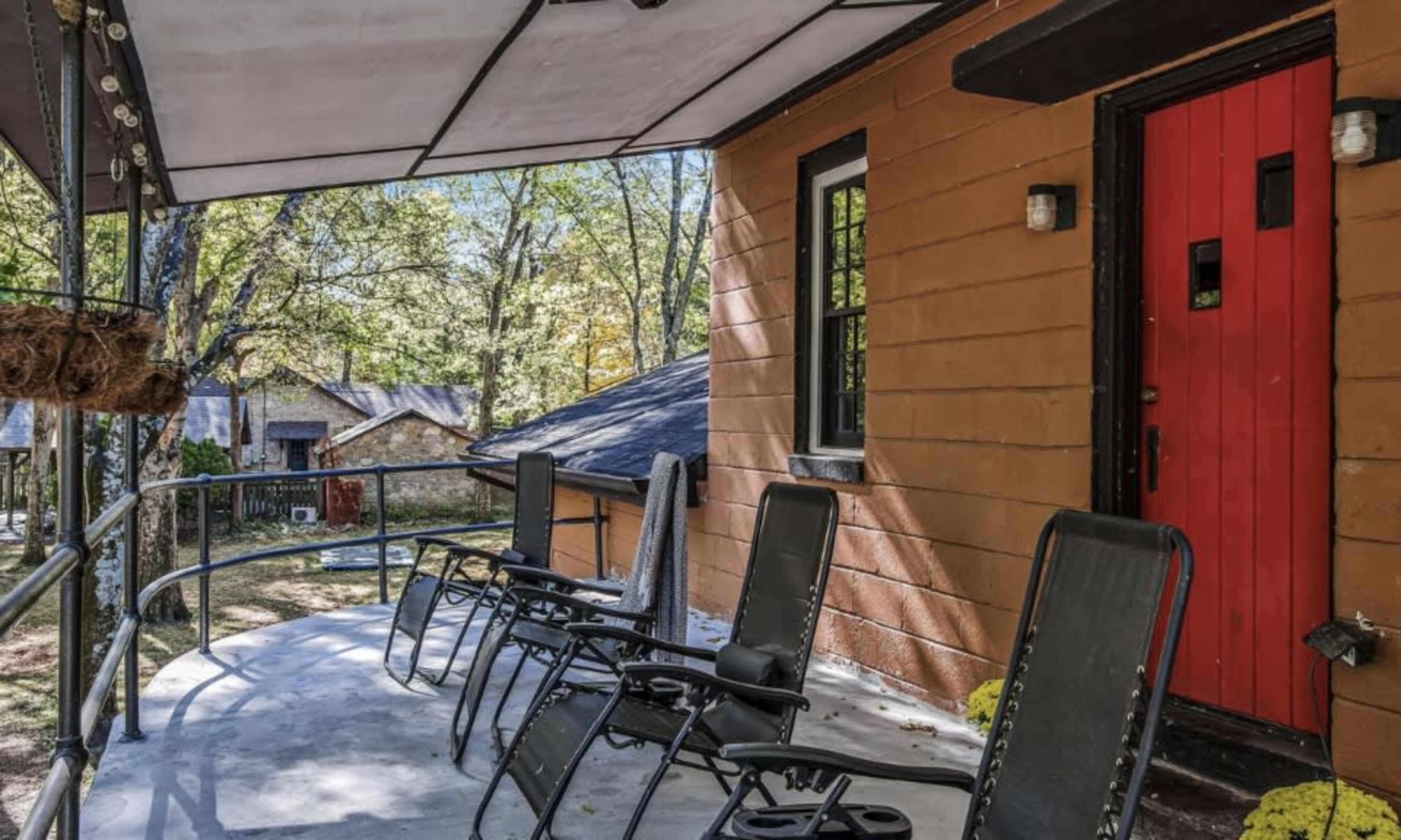 A porch with several black lounge chairs and a bright red front door, surrounded by trees and houses in the background.