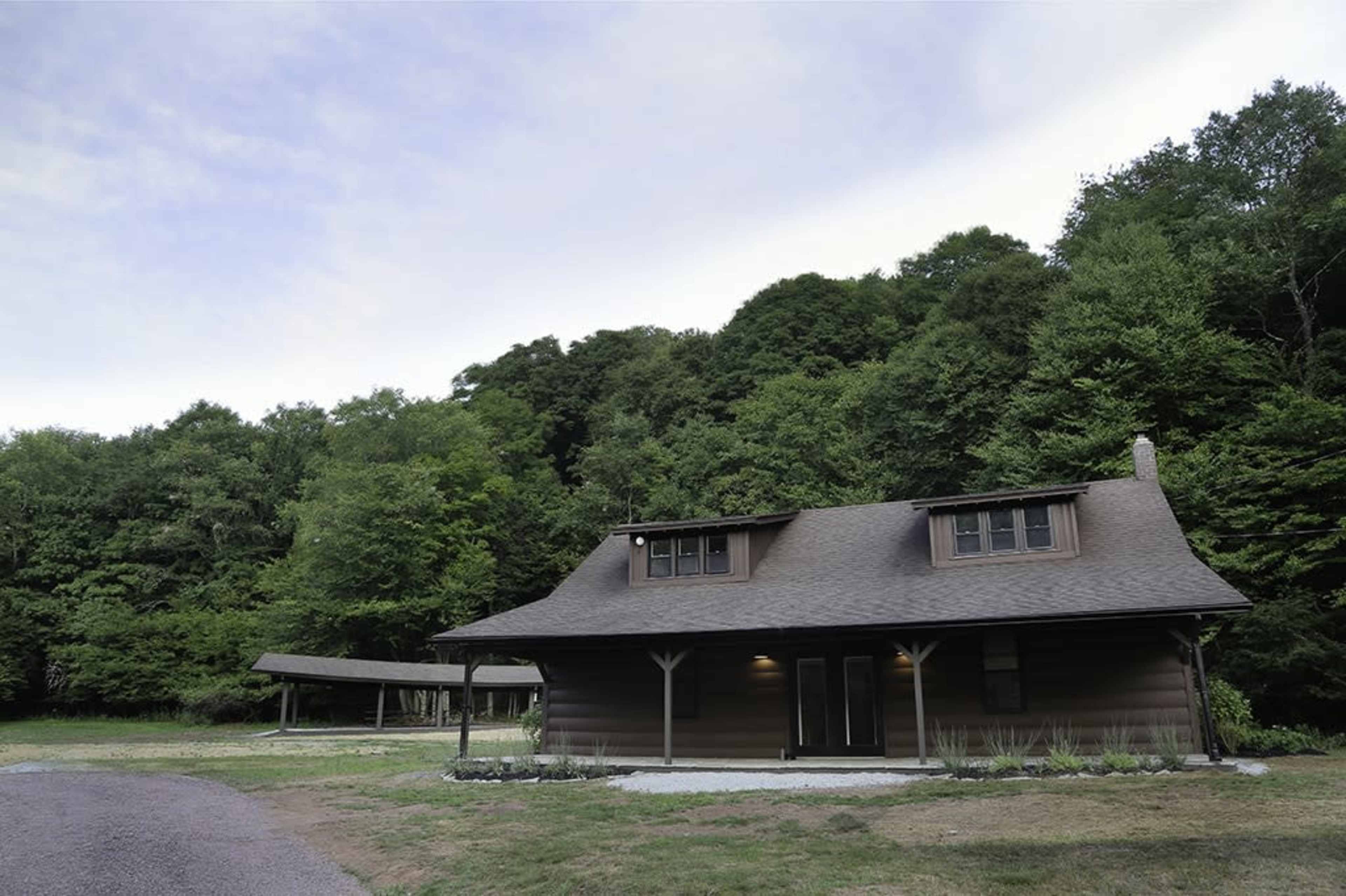 A cabin-style house is situated on a gravel driveway, surrounded by greenery and trees in the background.