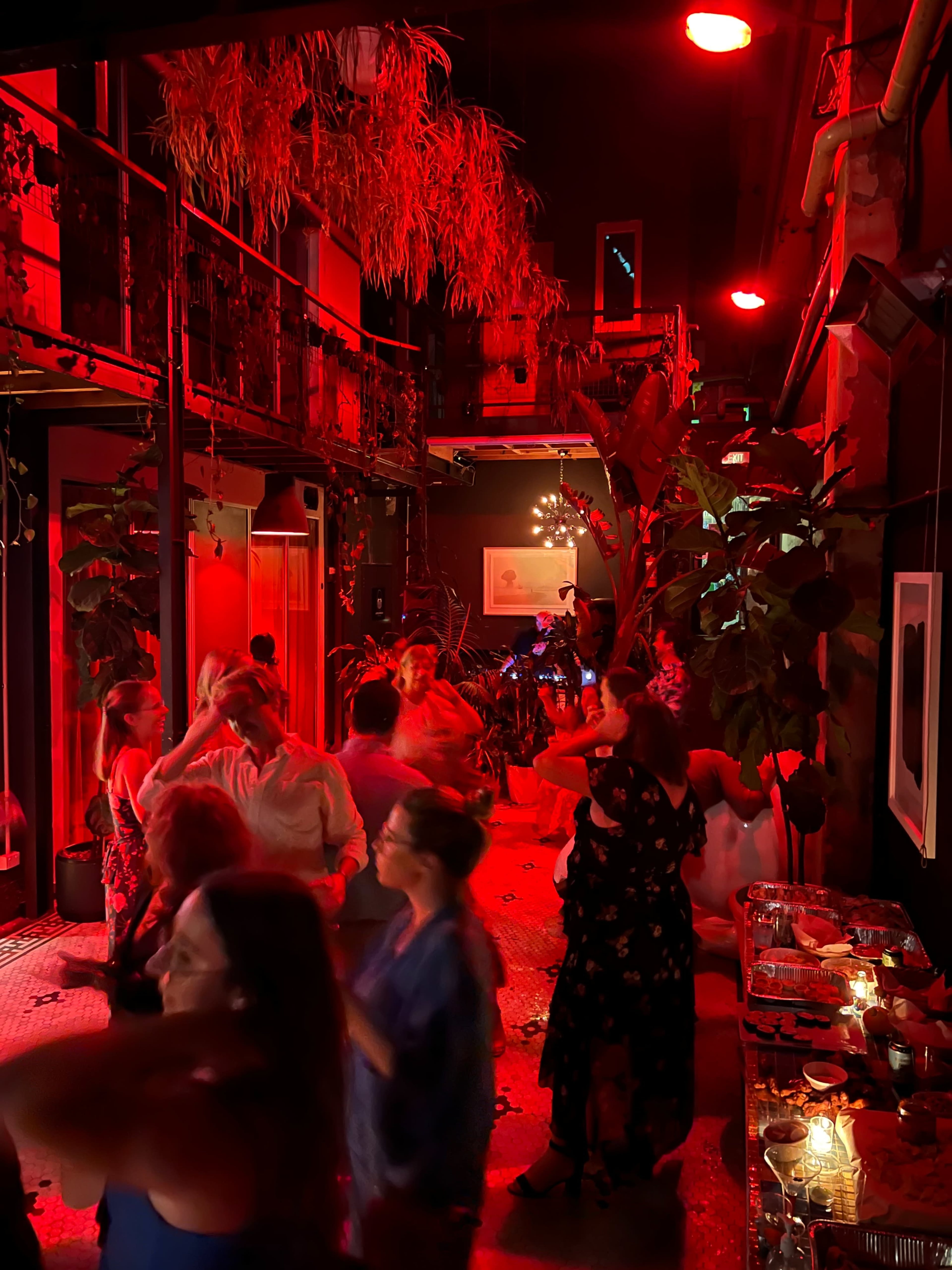 A lively party scene with people dancing under red lighting in a decorated indoor space filled with plants.