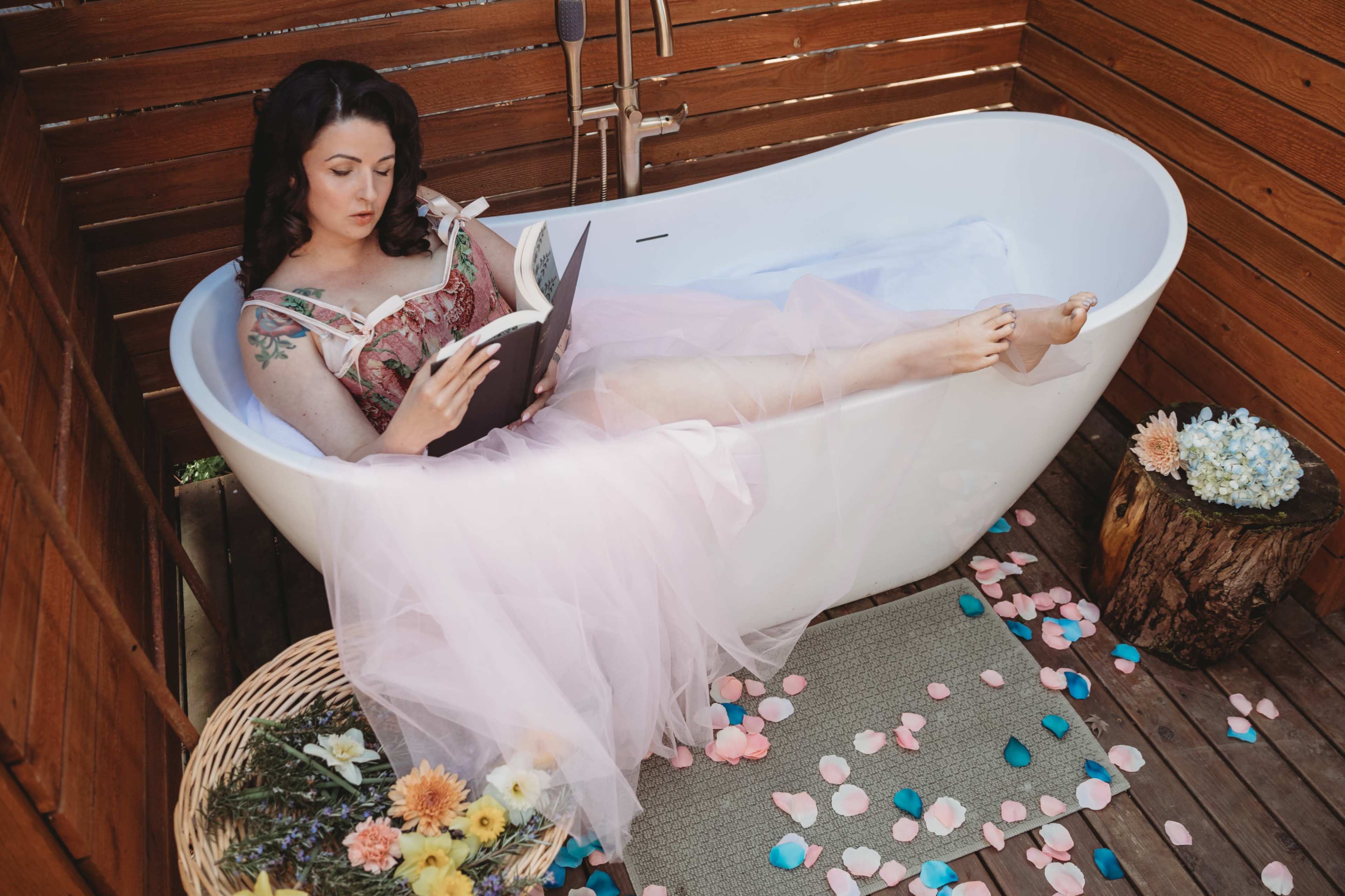 A woman reads a book while relaxing in a modern white bathtub surrounded by wooden walls and decorative flowers.