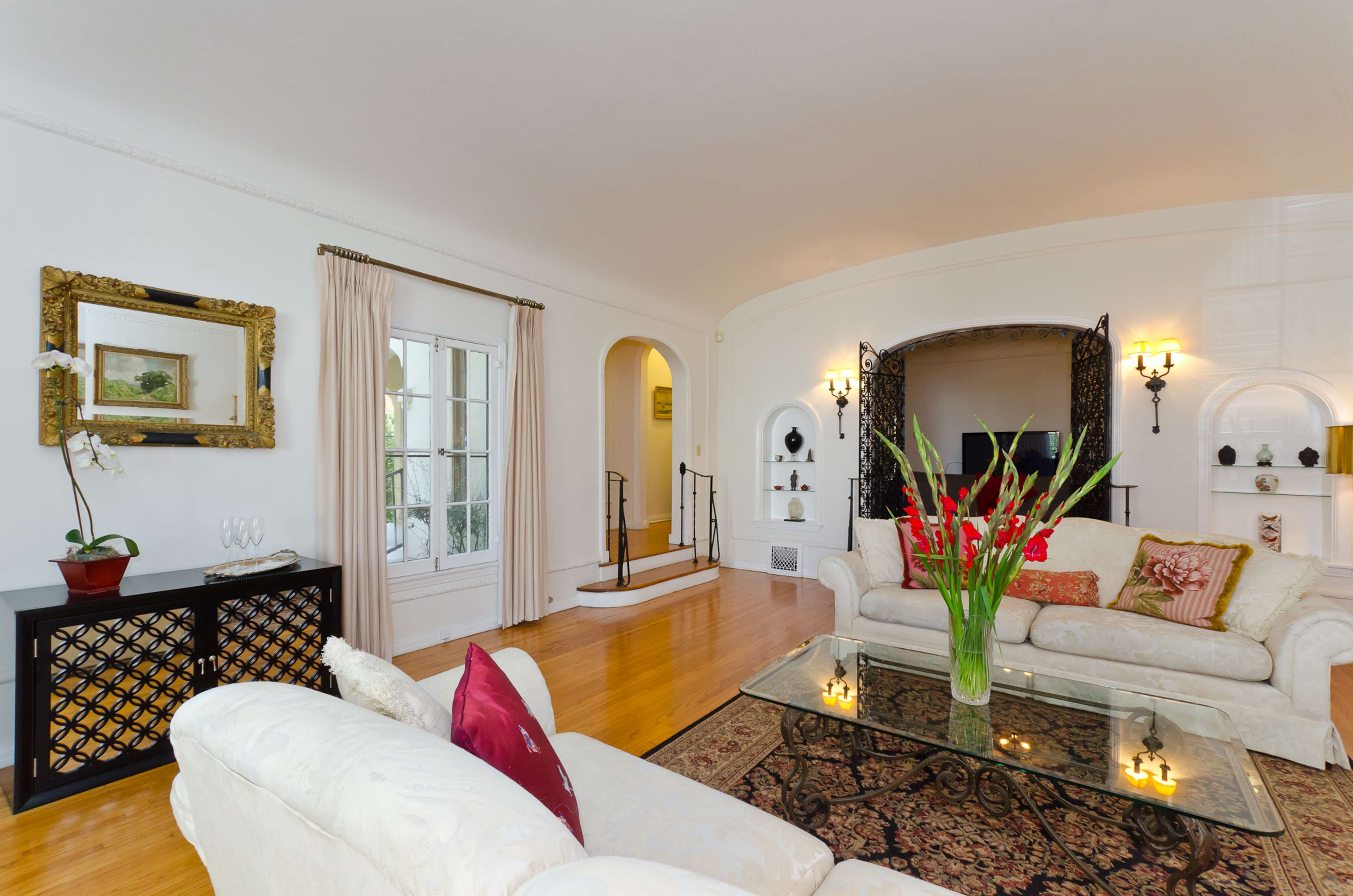 The scene features a bright living room with a white sofa, glass coffee table, and decorative plants, leading to an arched entryway with additional seating and wall-mounted shelves.