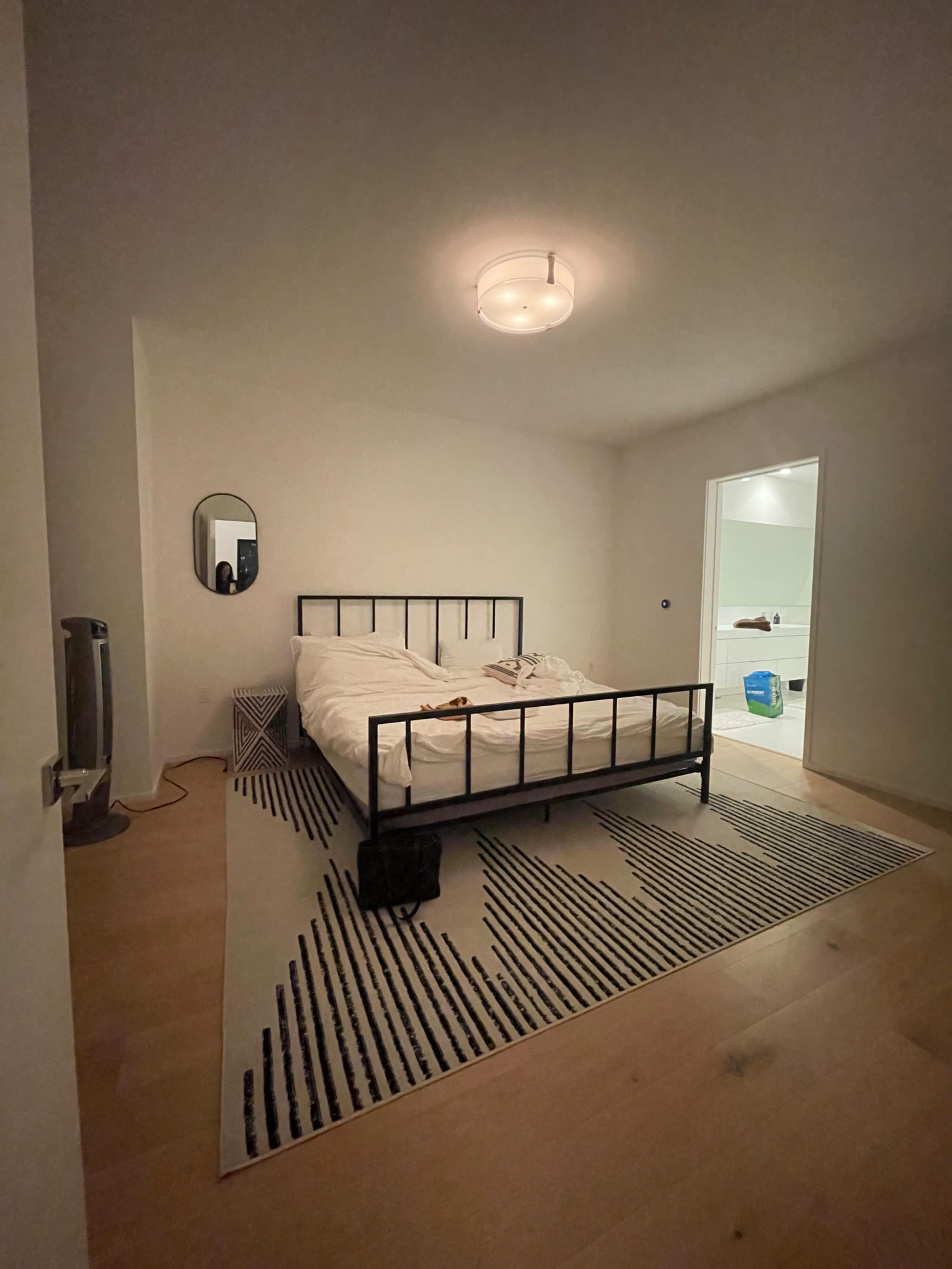 A bedroom features a bed with a white comforter, a round mirror on the wall, and a black and white patterned rug on the floor.