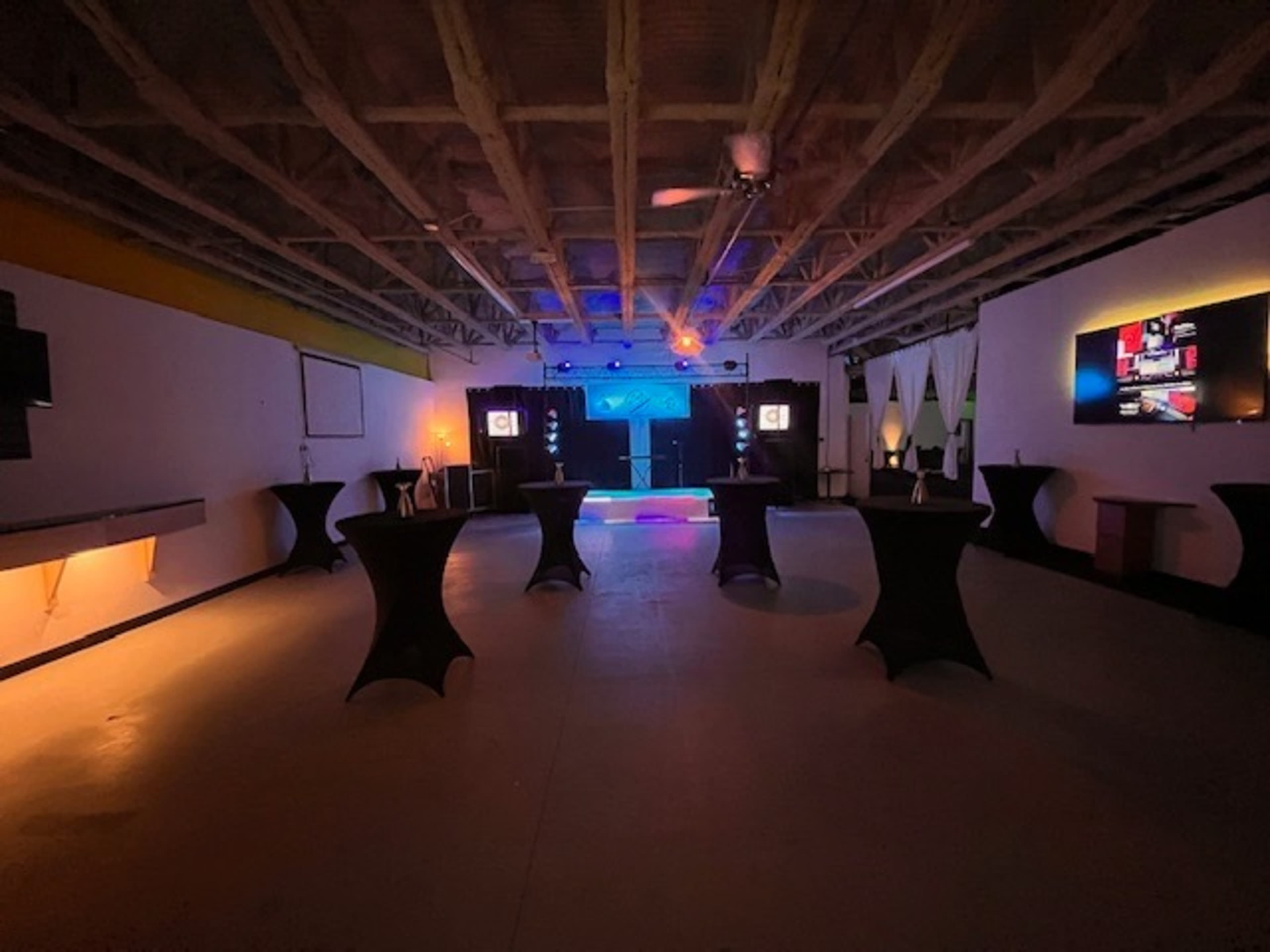 The image shows a large, empty event space with dark tables arranged around a central stage area lit by colorful lights.