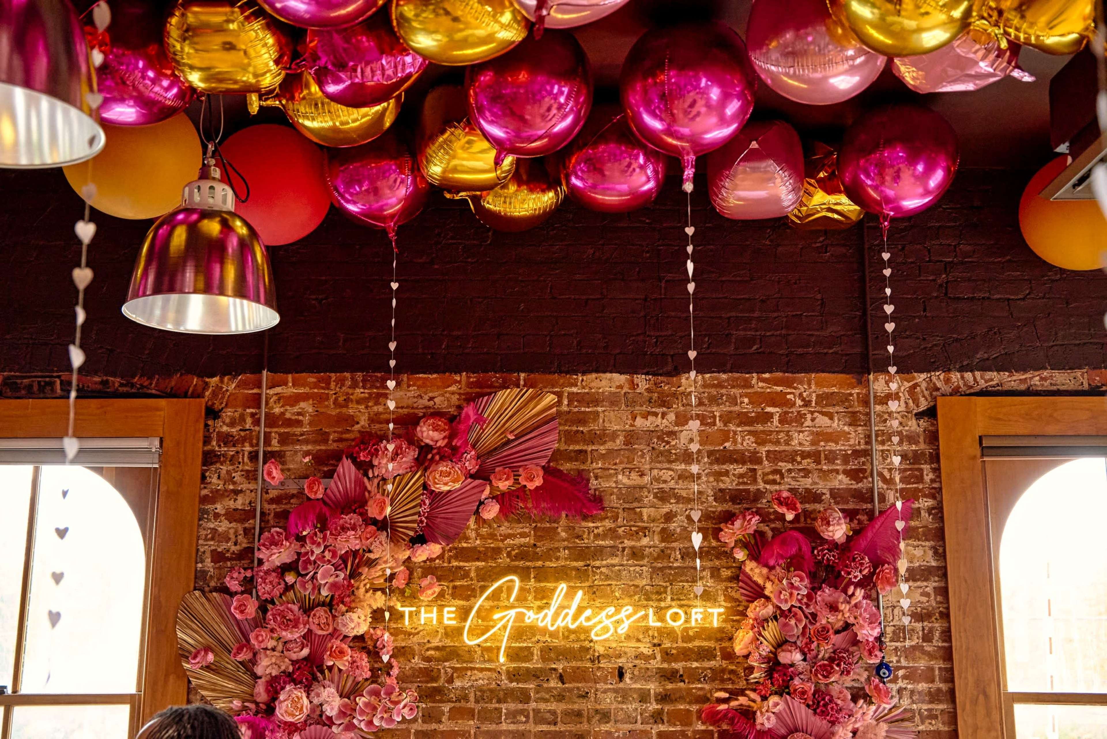 A decorated wall featuring floral arrangements and a neon sign that reads "THE Goddess LOFT," with colorful balloons above.
