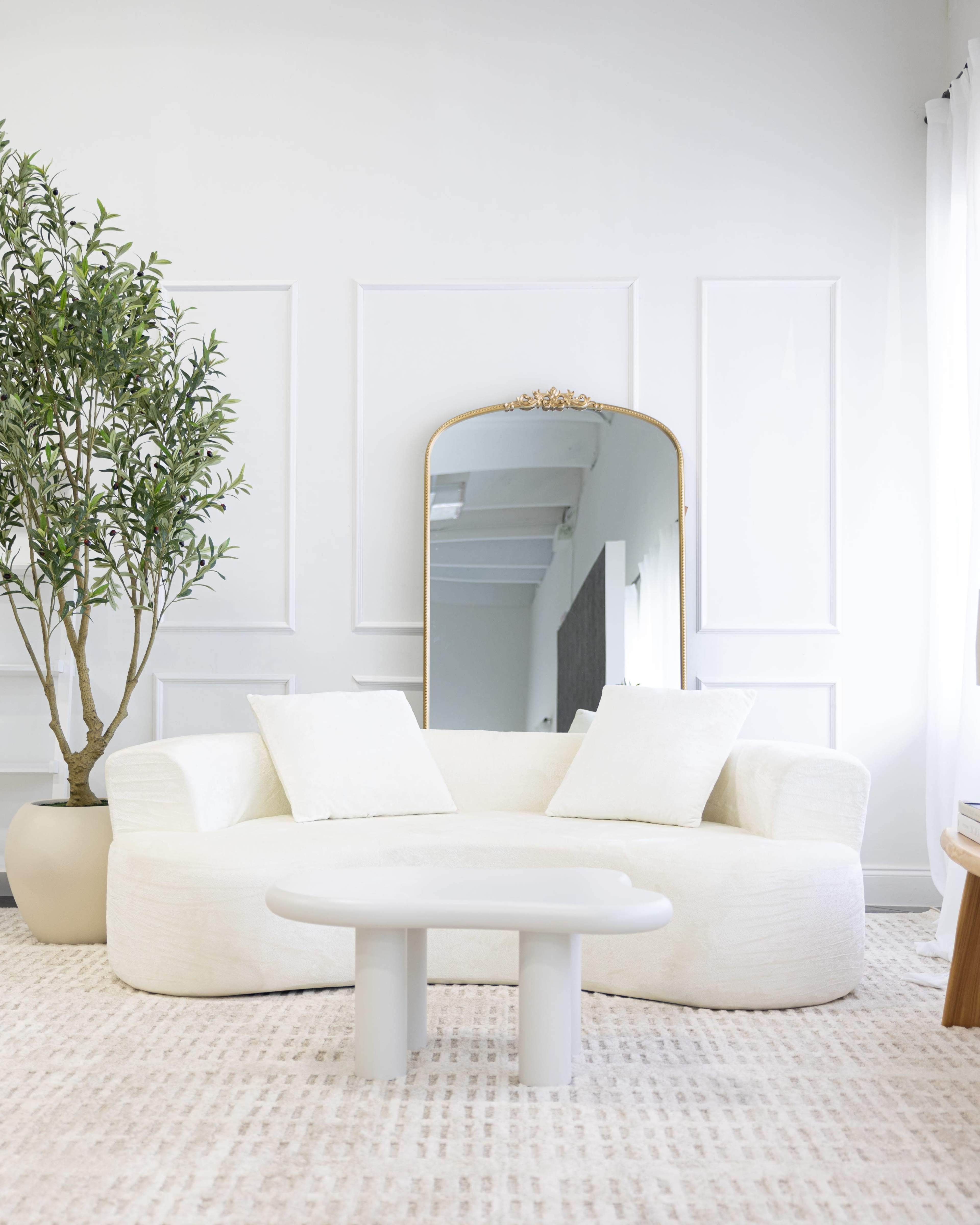 A white, curved couch with two cushions sits across from a round table in a modern living room, accompanied by a large mirror and a potted plant.