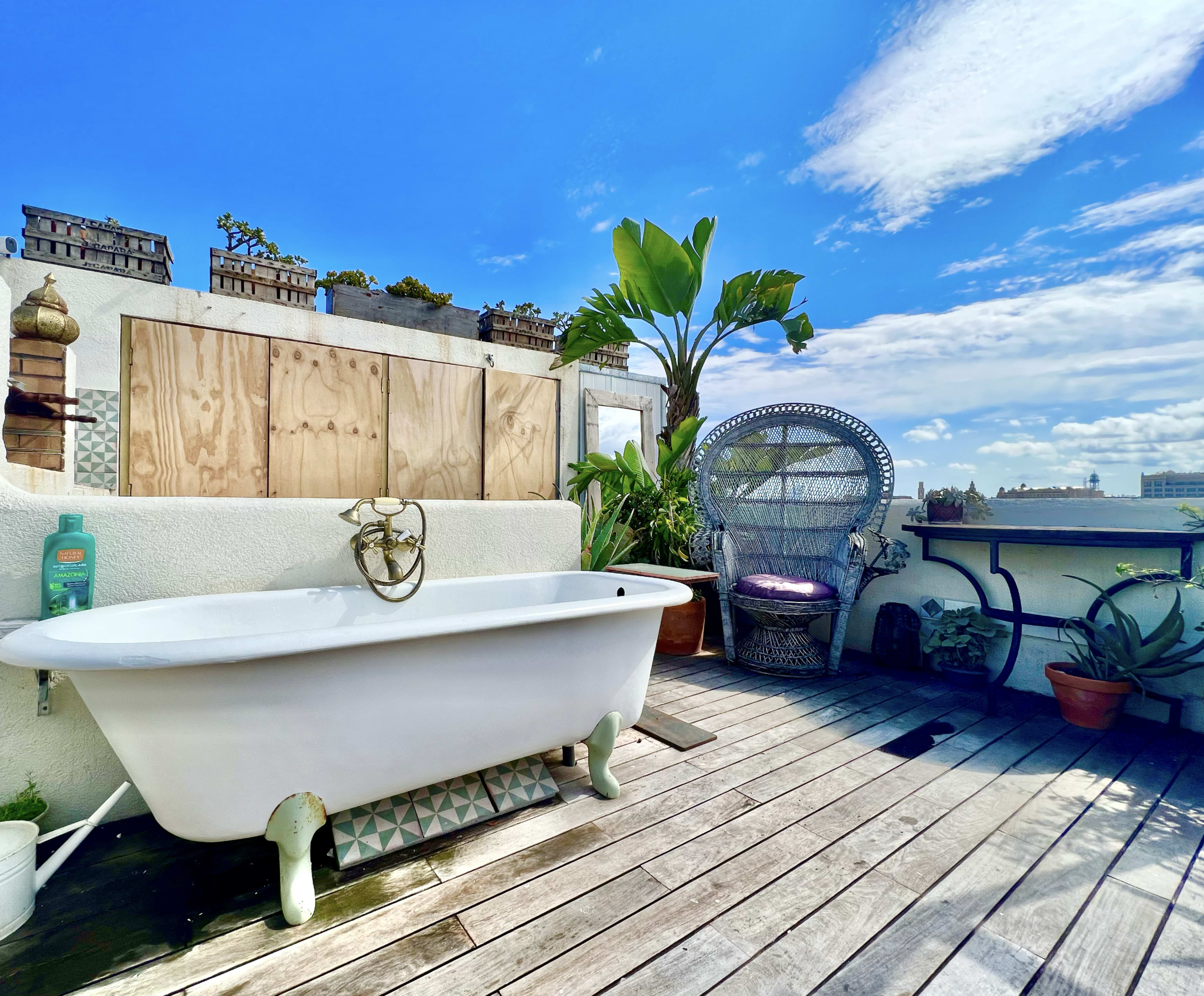 A vintage clawfoot bathtub is placed on a wooden deck surrounded by potted plants and a wicker chair under a blue sky with clouds.