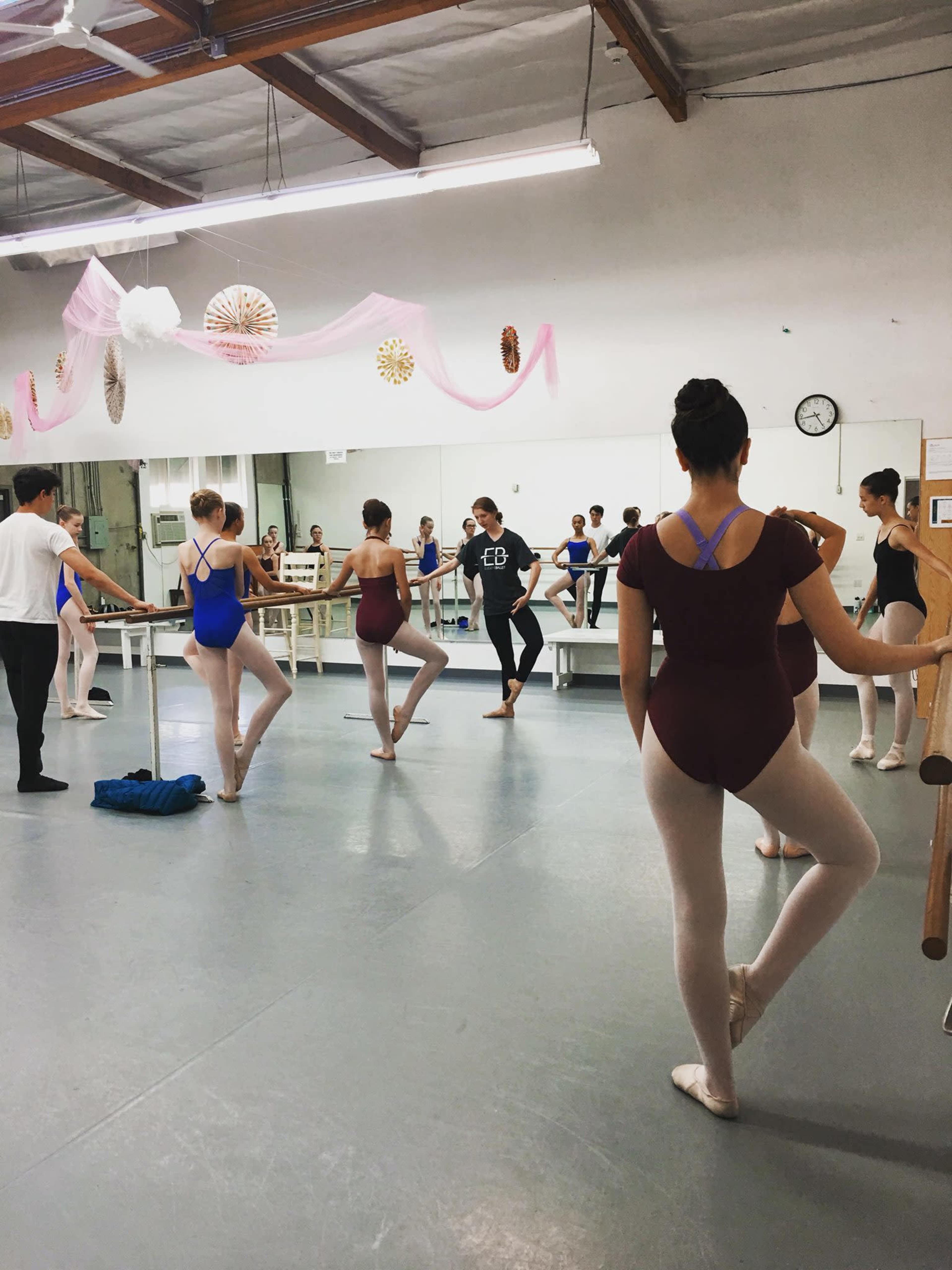 A group of ballet students practices at a dance studio with mirrors and barre equipment.